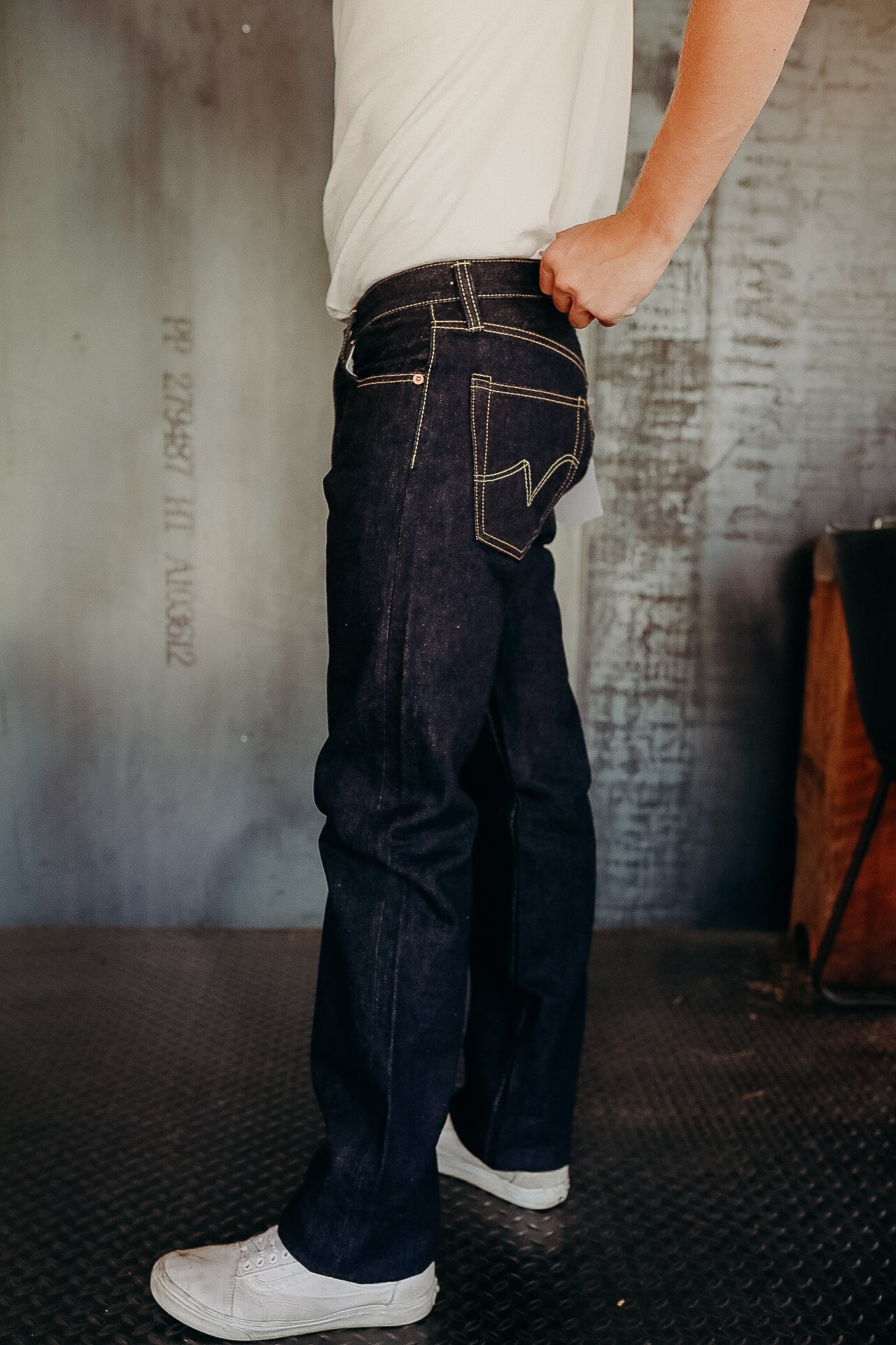 Person wearing dark raw denim jeans with yellow stitching and white sneakers in industrial setting