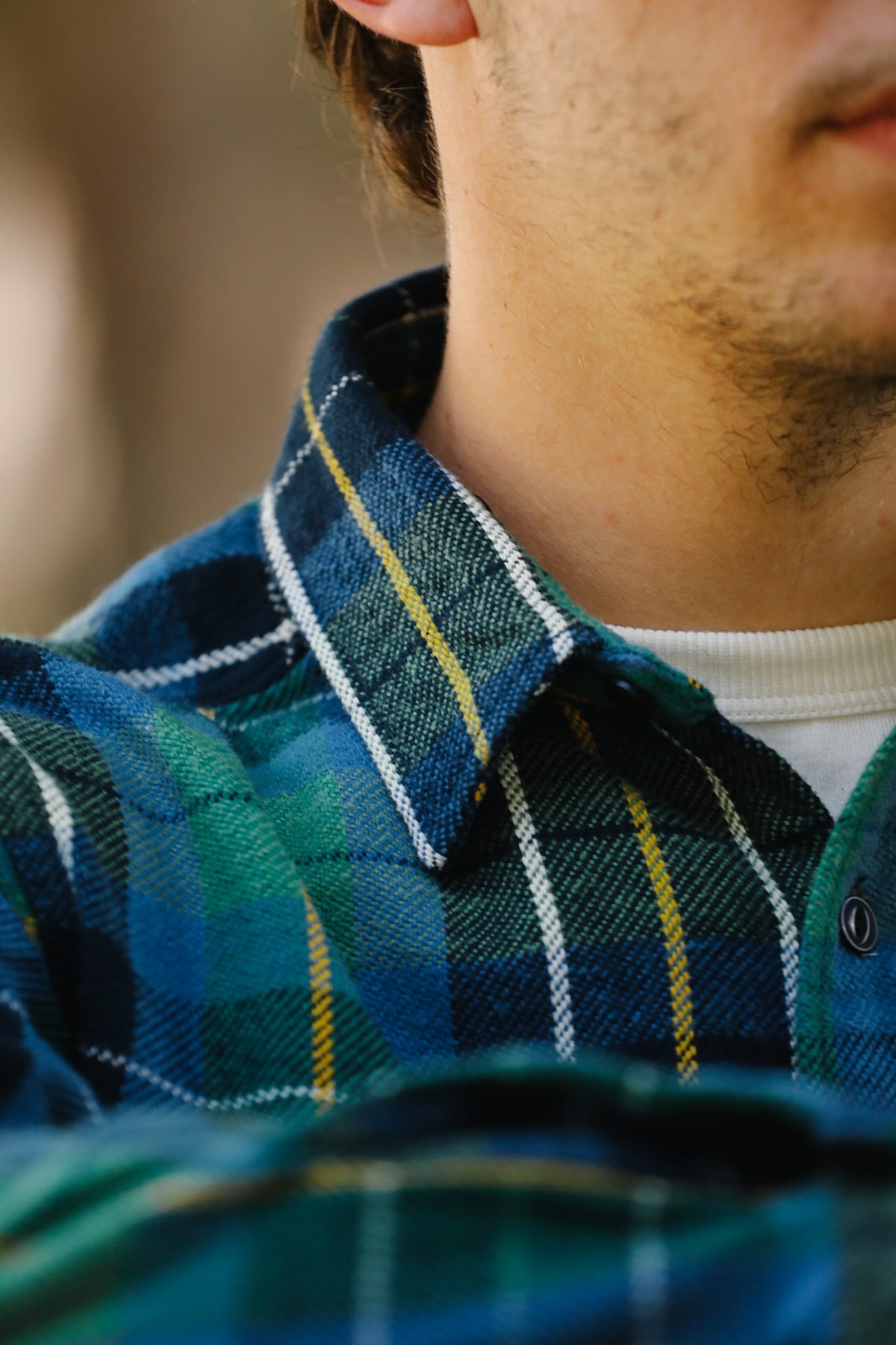 Close-up of a person wearing a blue and green plaid button-up shirt with thin yellow and white stripes, layered over a white undershirt.