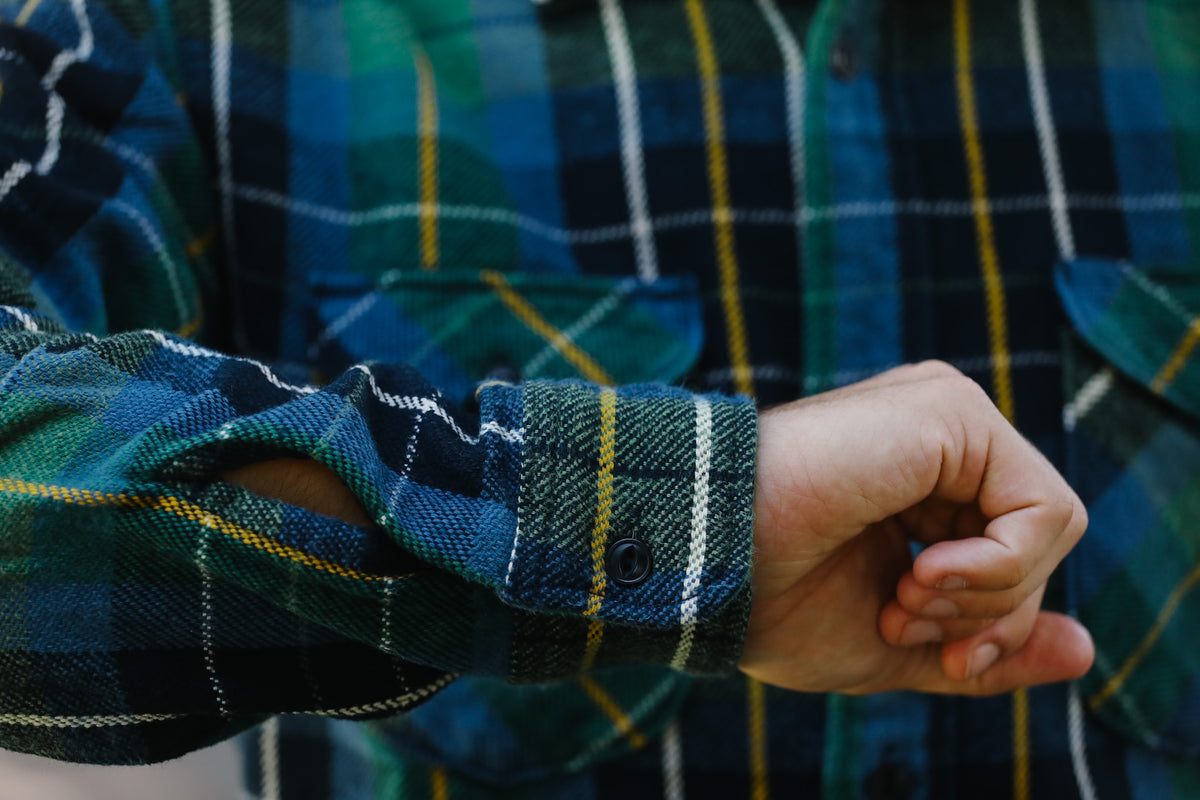 Close-up of a person wearing a green and blue plaid flannel shirt, showing the rolled-up cuff and hand.