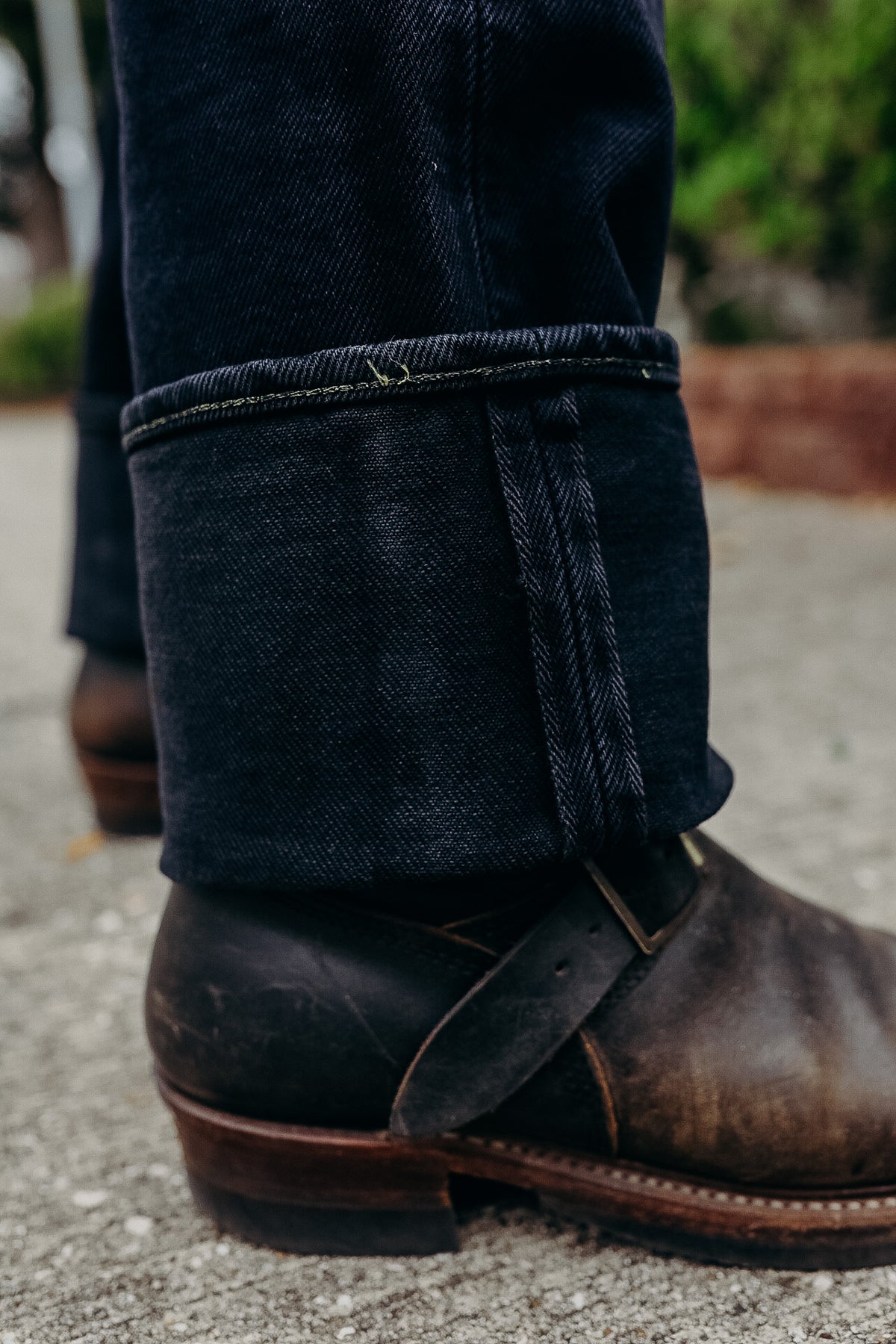 Close-up of cuffed dark denim jeans paired with worn brown leather boots on concrete surface