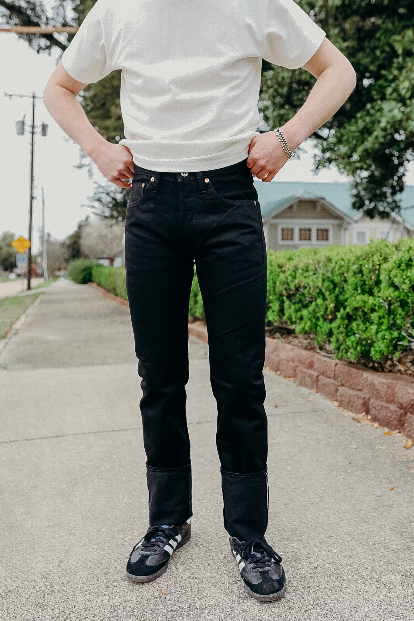 Person wearing black jeans with cuffs and black sneakers with white stripes on sidewalk