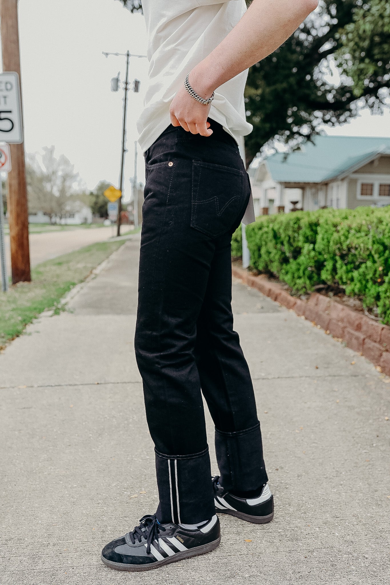 Person wearing black denim jeans with cuffed hems and black Adidas sneakers on sidewalk