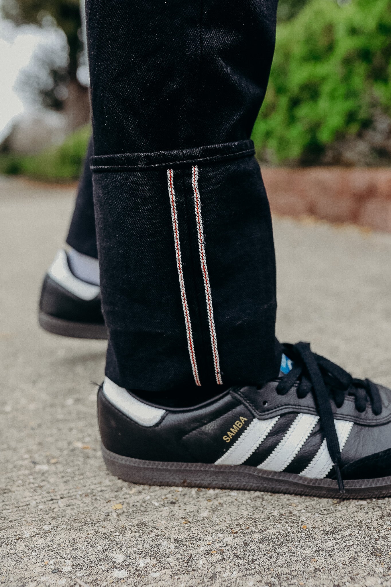 Close-up of black denim jeans with white and red selvedge stitching alongside black Adidas Samba sneakers on concrete