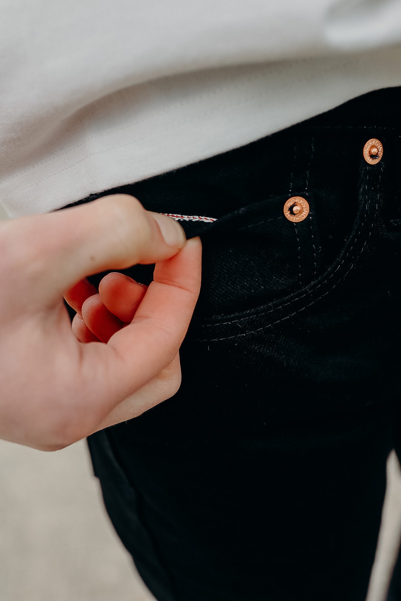Hand holding the edge of a black denim jean pocket with copper rivets and white fabric background