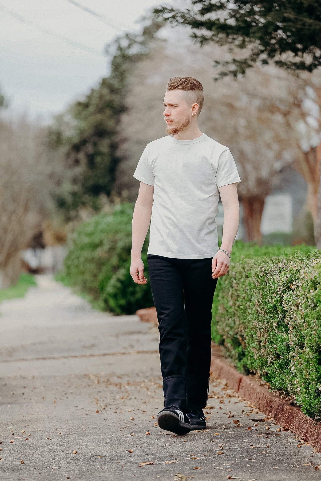 Man walking on sidewalk wearing white t-shirt and black pants with green bushes and trees outdoors