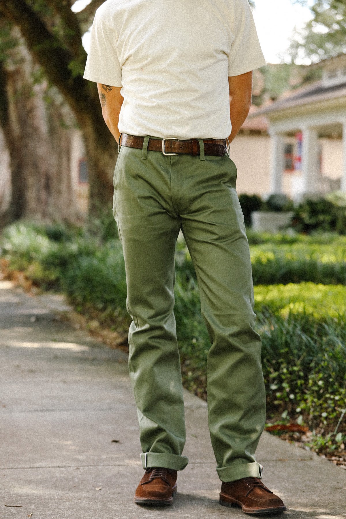 A man standing outdoors on a sidewalk wearing olive green chino pants, a white t-shirt, a brown leather belt, and brown leather shoes.