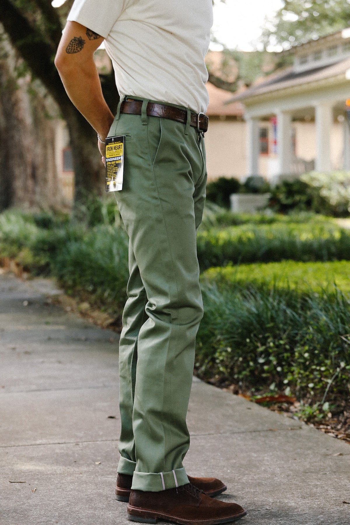 A person standing outdoors on a sidewalk wearing olive green trousers, a white shirt, a dark brown belt, and brown shoes. A small yellow-and-black hangtag is attached near the pocket area of the pants.