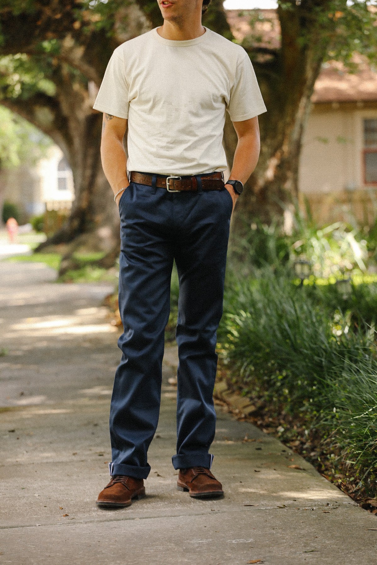 A man standing on an outdoor sidewalk wearing a cream t-shirt, navy blue pants, a brown leather belt, and brown leather shoes.