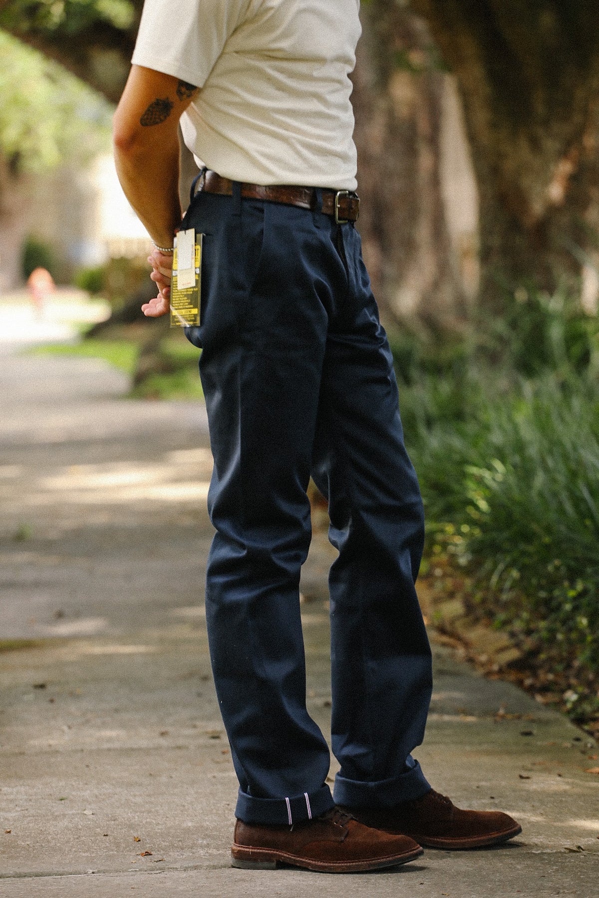 A man wearing dark navy blue chino pants, a white t-shirt, and brown leather shoes, standing on a tree-lined pathway and holding a glass of beer.