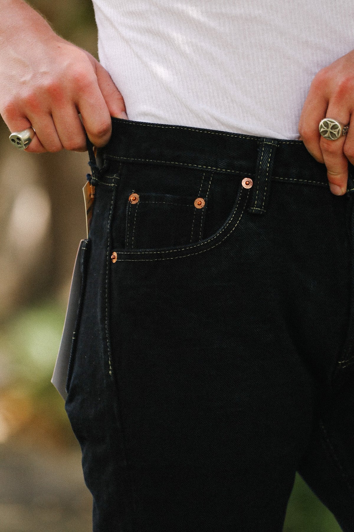 Close-up of a person wearing dark indigo denim jeans with copper-toned rivets and a white shirt, pulling the waistband slightly outward with both hands.