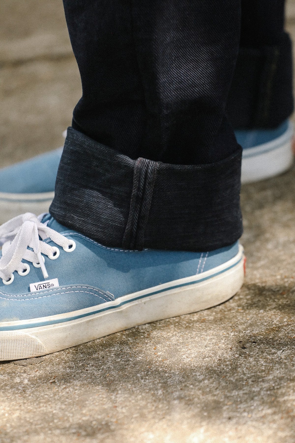 A close-up of a person's feet wearing light blue Vans sneakers with dark cuffed jeans, standing on a textured concrete ground.