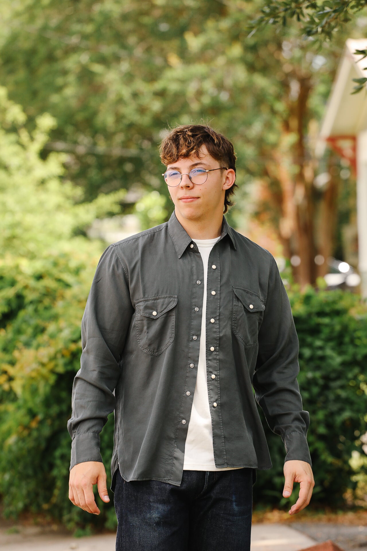 Young man wearing a dark gray button-up shirt with chest pockets and glasses outdoors in a green leafy setting