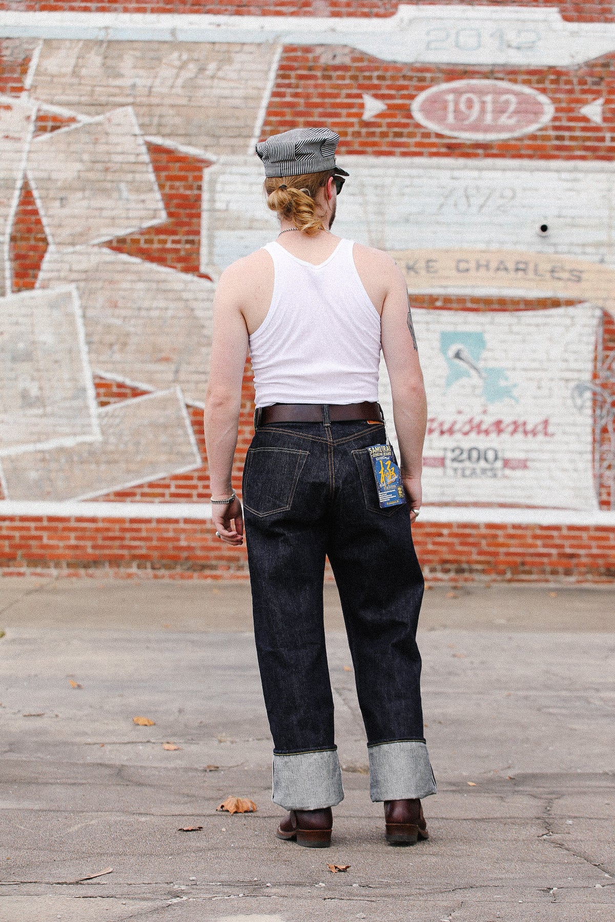 A person standing outdoors with their back to the camera, wearing a white tank top, dark blue rolled-calf denim jeans, a brown belt, and a gray cap, in front of a brick wall with painted graffiti.