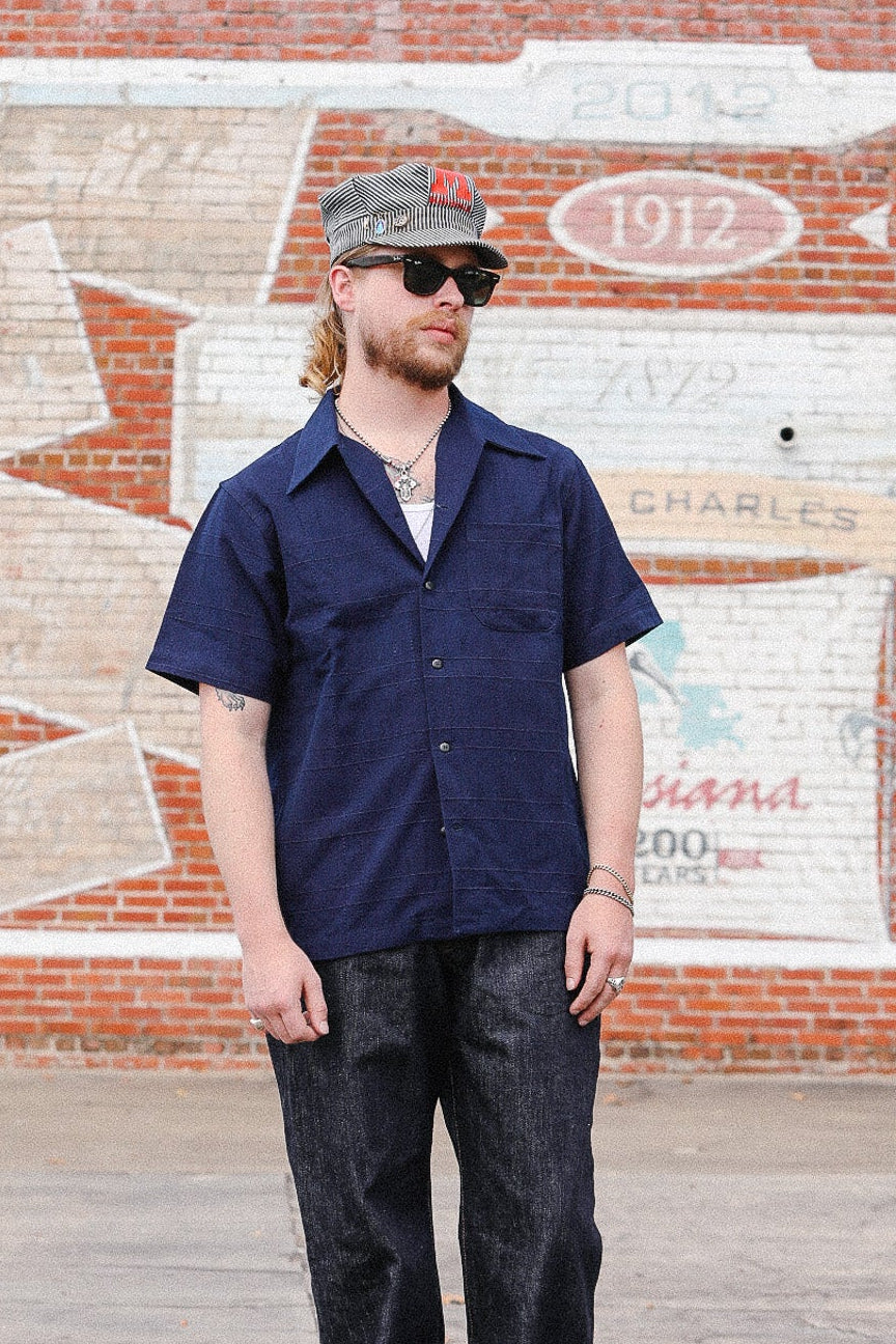 A man wearing a navy blue short-sleeve button-up shirt, dark jeans with rolled cuffs, brown shoes, and a baseball cap stands in front of a brick wall with a painted mural.