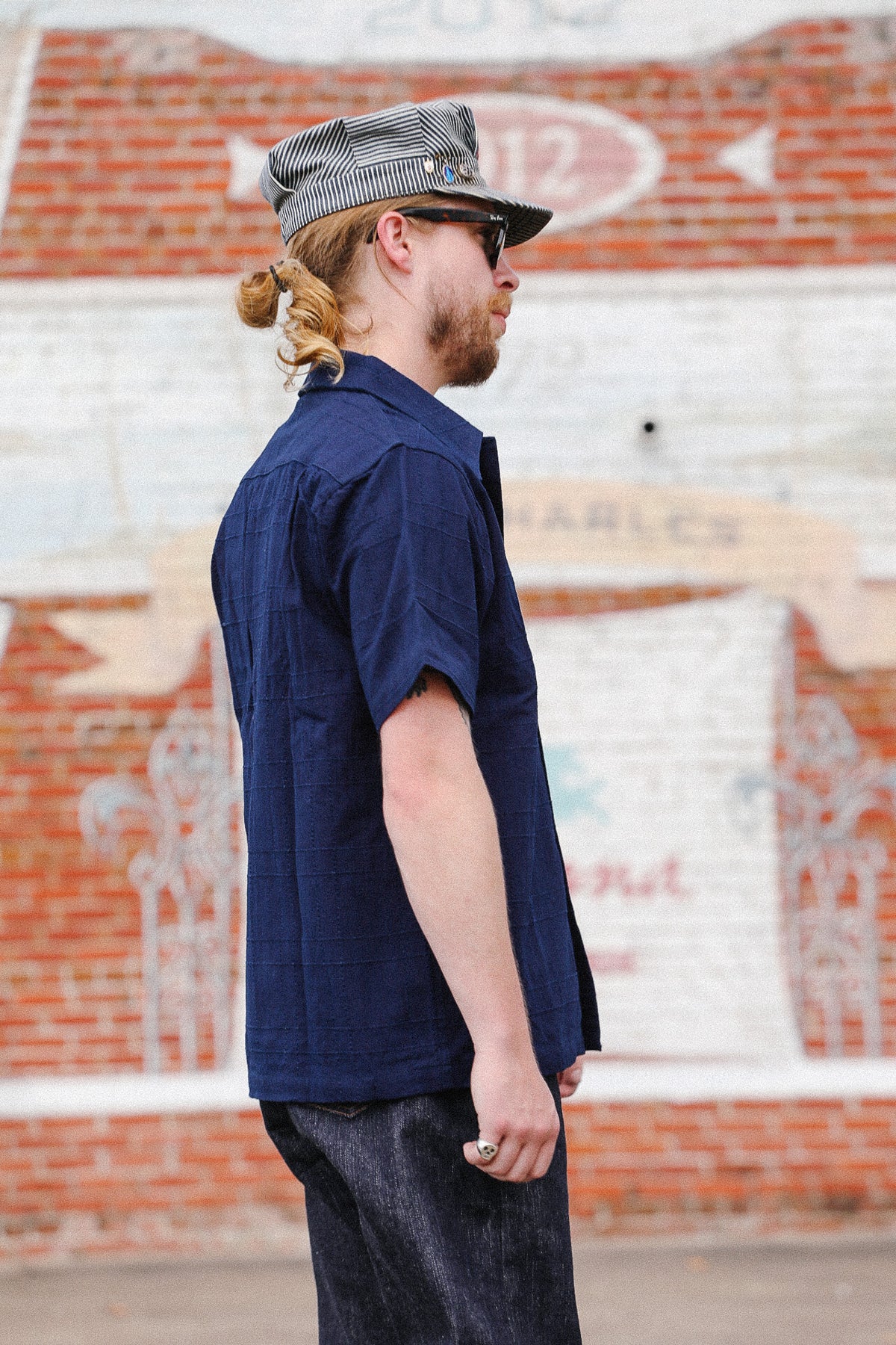 A man wearing a navy blue short-sleeve button-up shirt, dark jeans, a gray and white striped cap, and sunglasses, standing in front of a brick wall with white painted lettering.