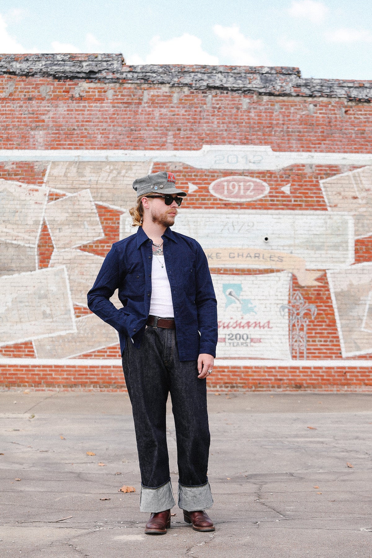 A man wearing a navy blue button-up shirt, dark jeans, brown boots, and a gray cap stands in front of a brick wall with a painted mural.
