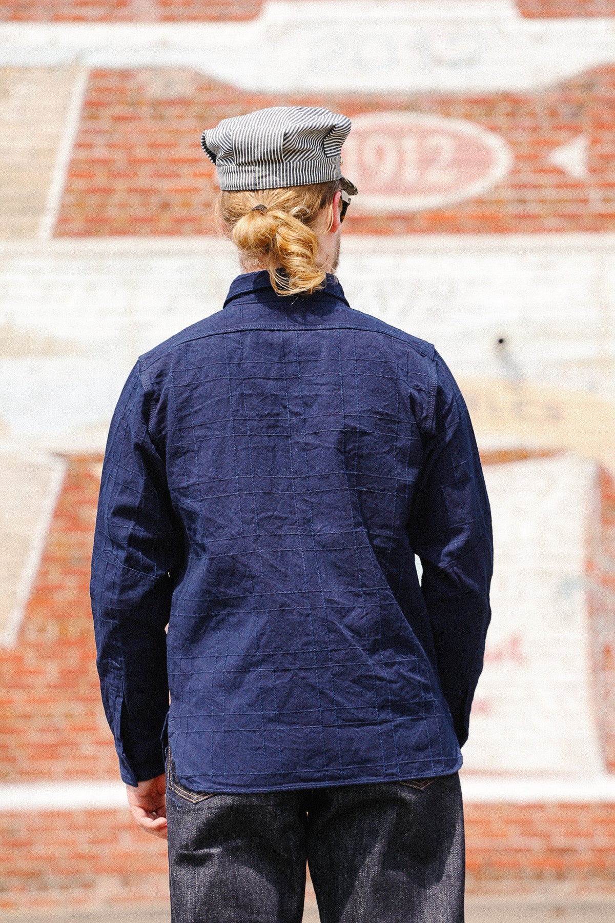 A person viewed from behind wearing a navy blue textured long-sleeve shirt and a gray striped flat cap, standing in front of a brick wall.