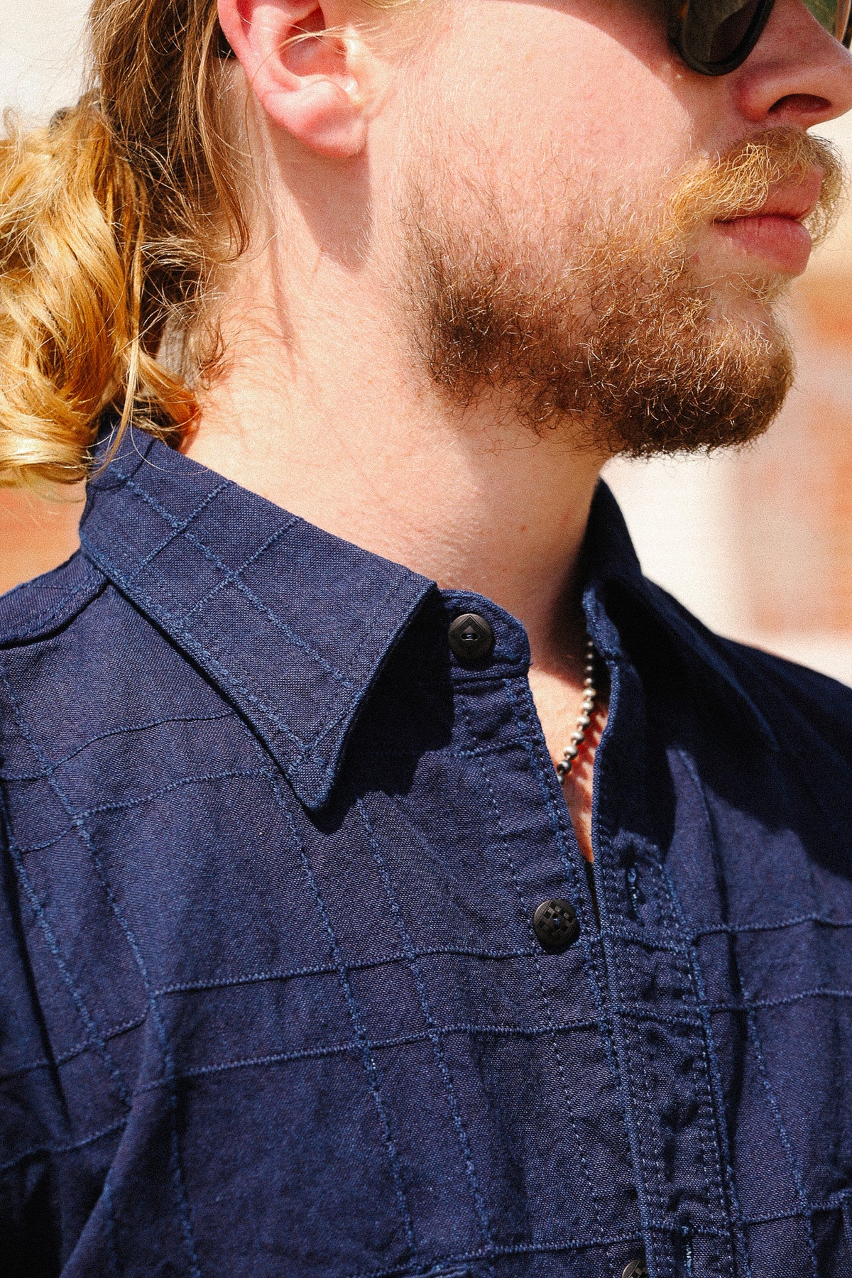 Close-up of a man wearing a dark navy blue textured button-down shirt, with a beard, sunglasses, and a ponytail visible.