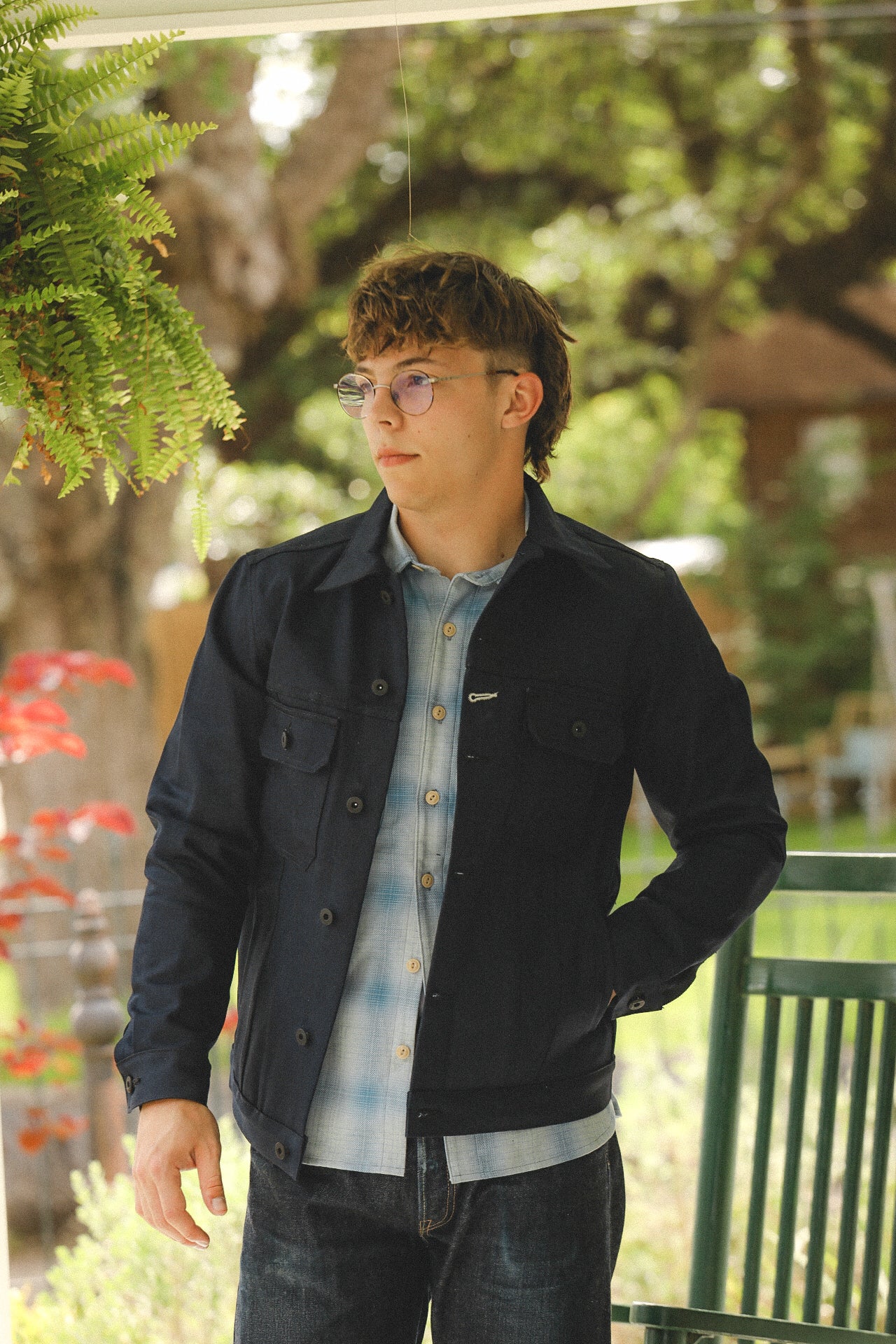 Young man wearing black jacket and plaid shirt standing outdoors under green trees with hanging fern