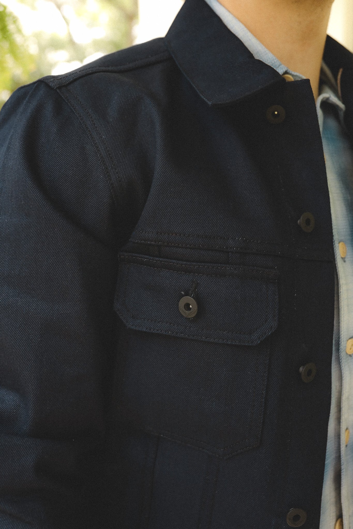 Close-up of a person wearing a dark blue denim jacket with buttoned chest pocket and light blue shirt underneath
