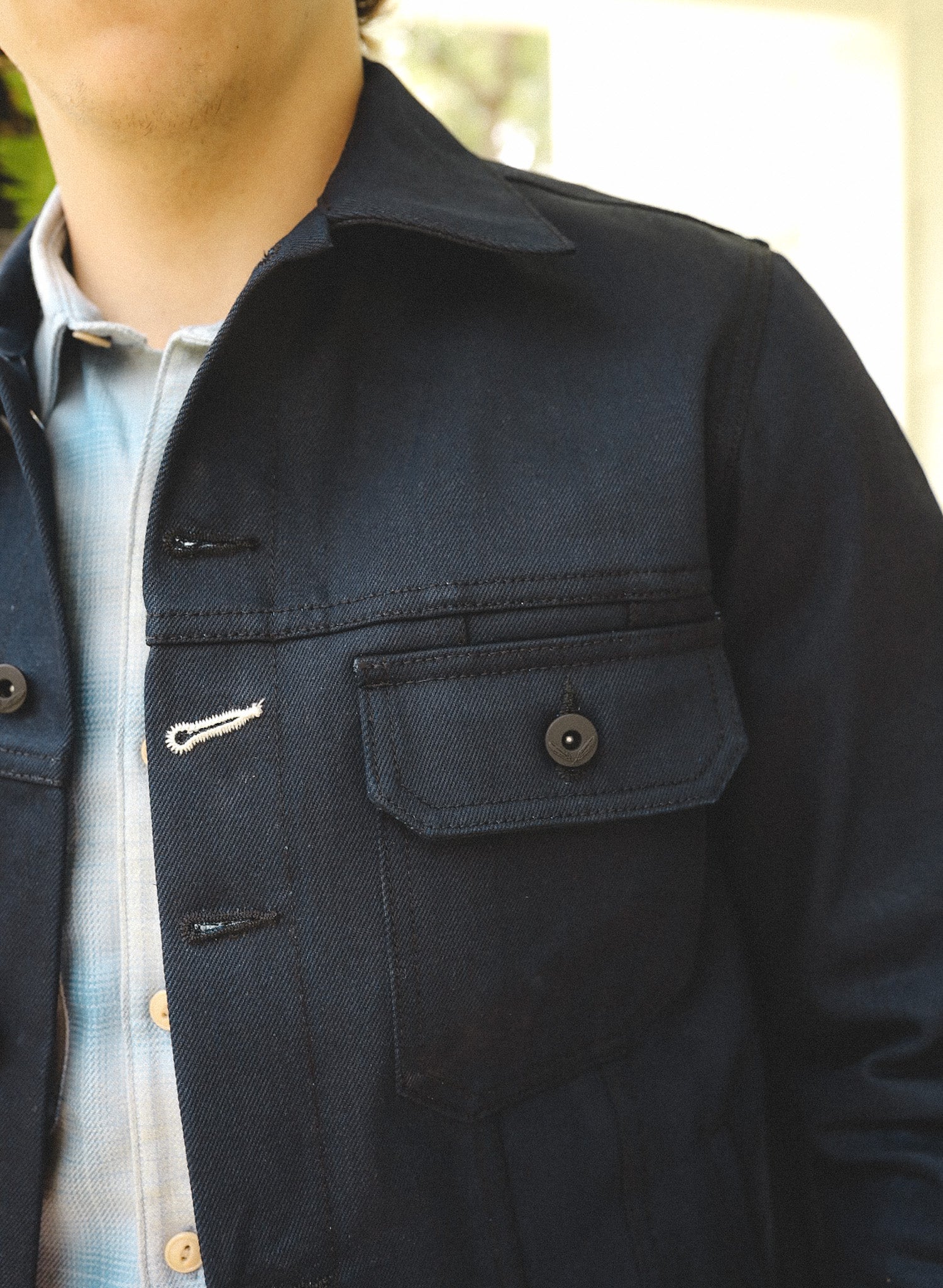 Close-up of a man wearing a dark navy work jacket with flap pockets over a light blue plaid shirt