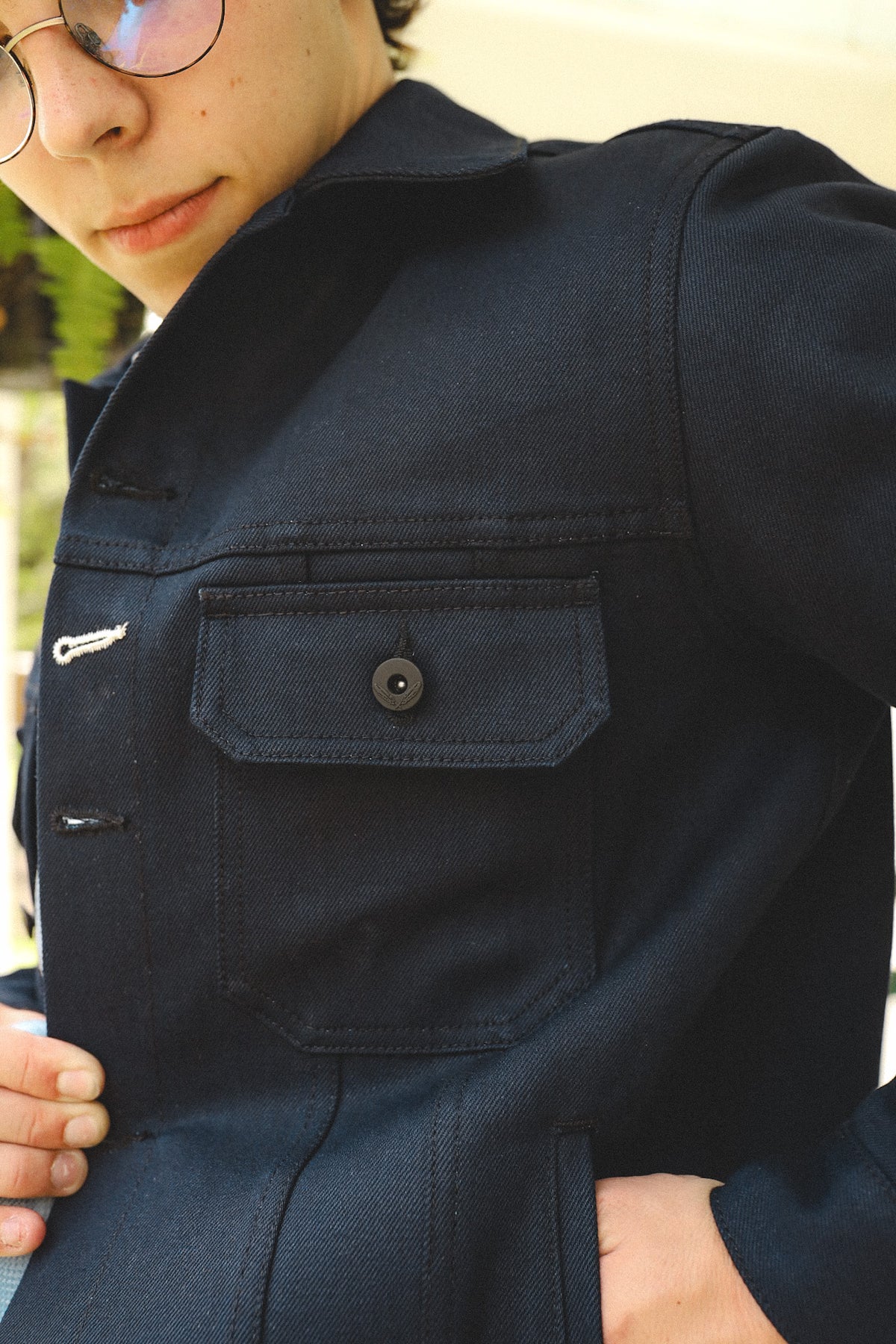 Close-up of person wearing dark denim jacket with buttoned chest pocket and glasses outdoors