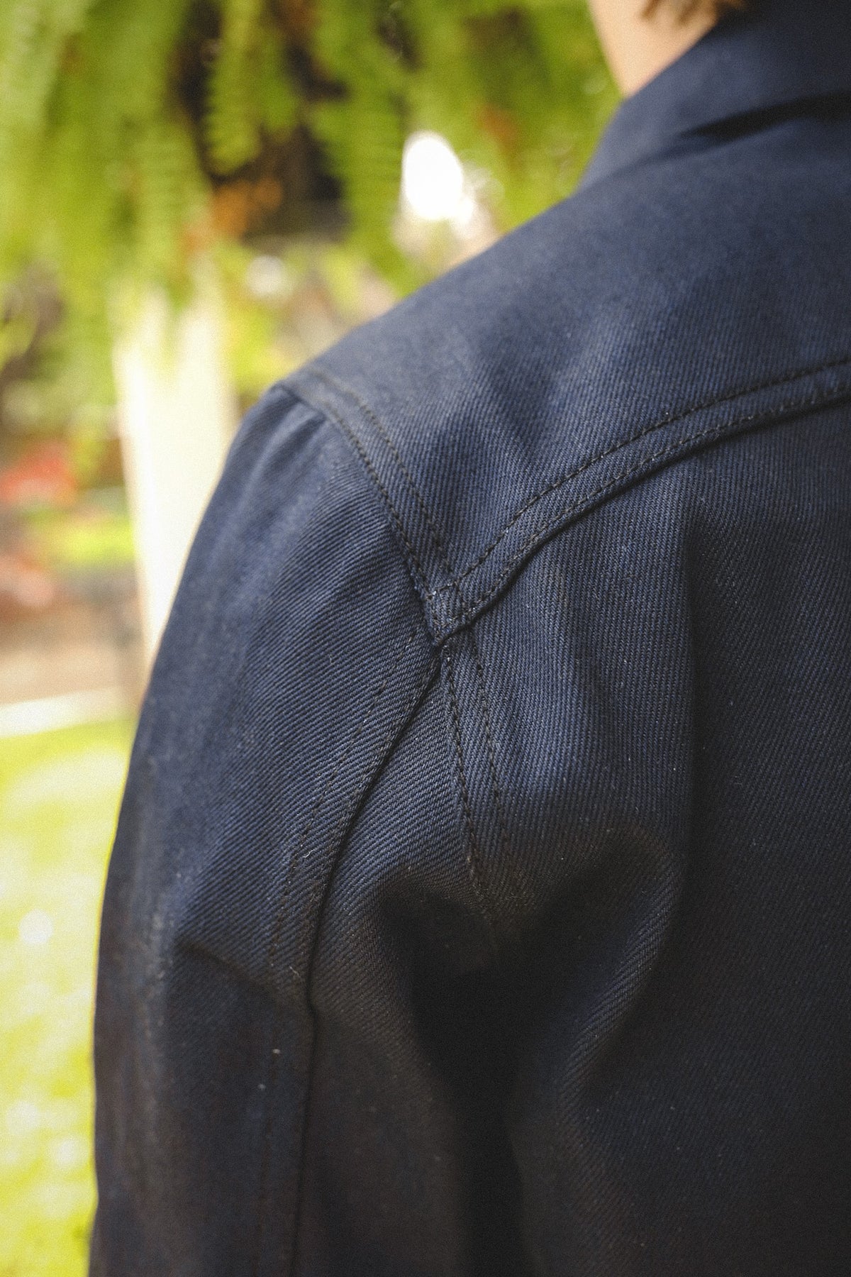 Close-up of shoulder seam detail on dark denim jacket with natural outdoor background