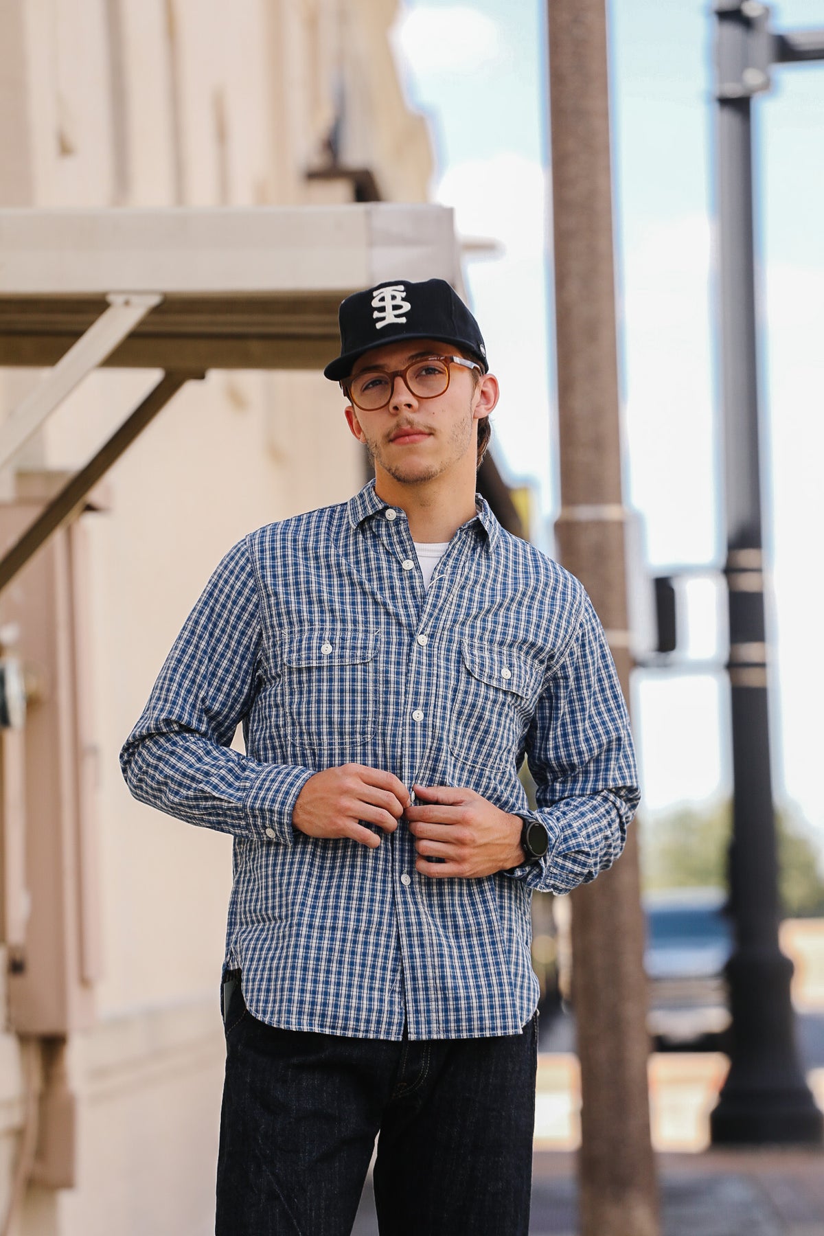 A man wearing a blue and white gingham long-sleeve button-down shirt, dark jeans, and a black baseball cap with a white logo, standing outdoors on a city street.