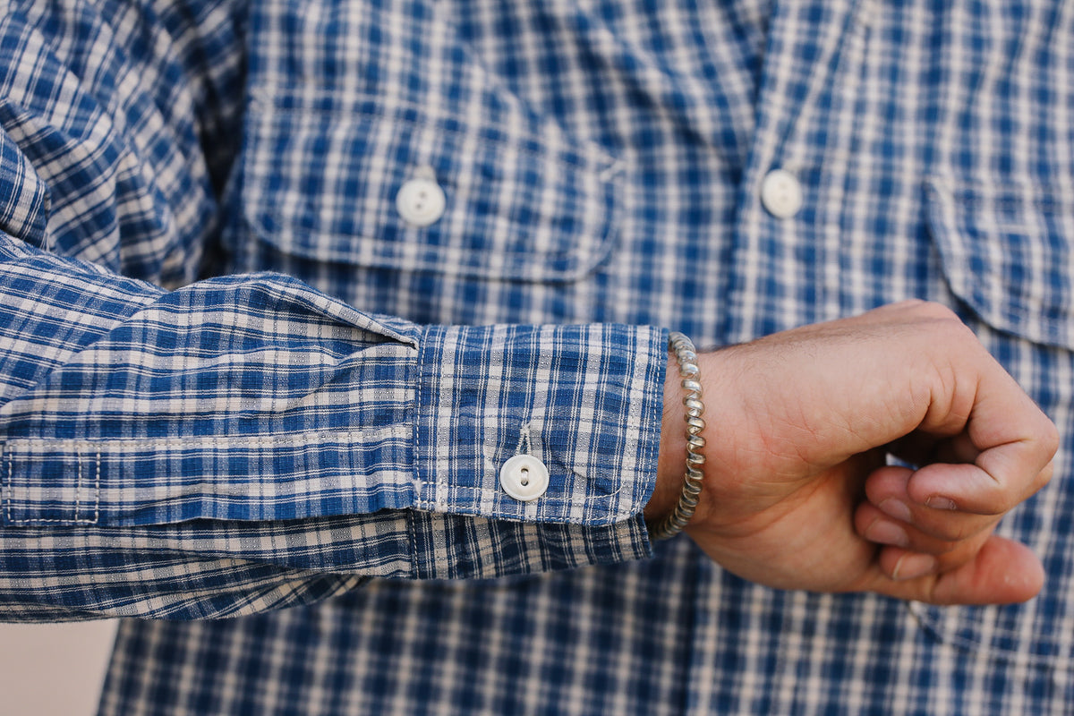 A close-up of a person's wrist wearing a silver beaded bracelet, showing the cuff of a blue and white plaid button-down shirt.
