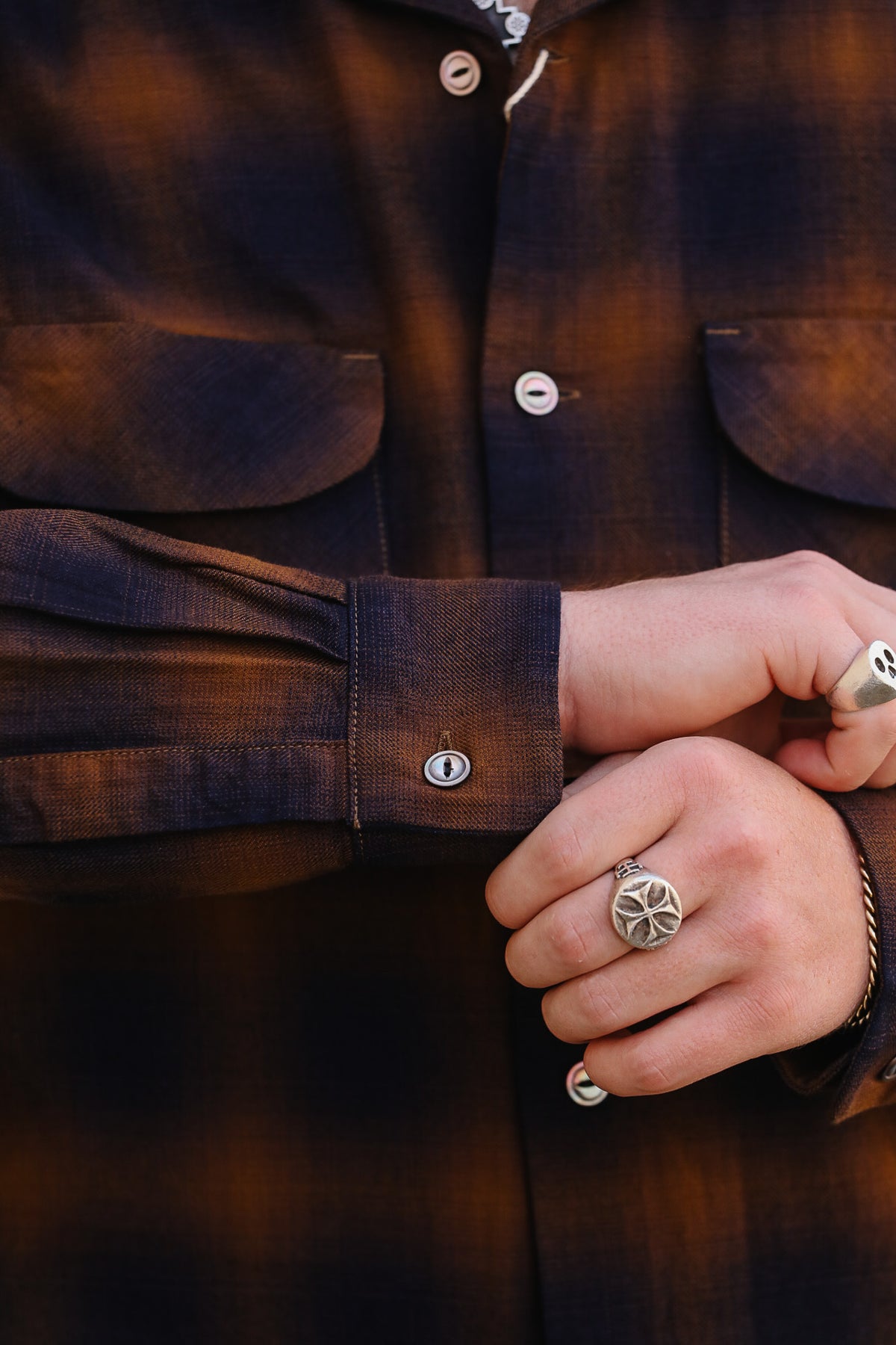 Close-up of a person wearing a dark brown and black plaid button-up shirt, with hands adjusting the cuff and wearing silver rings.