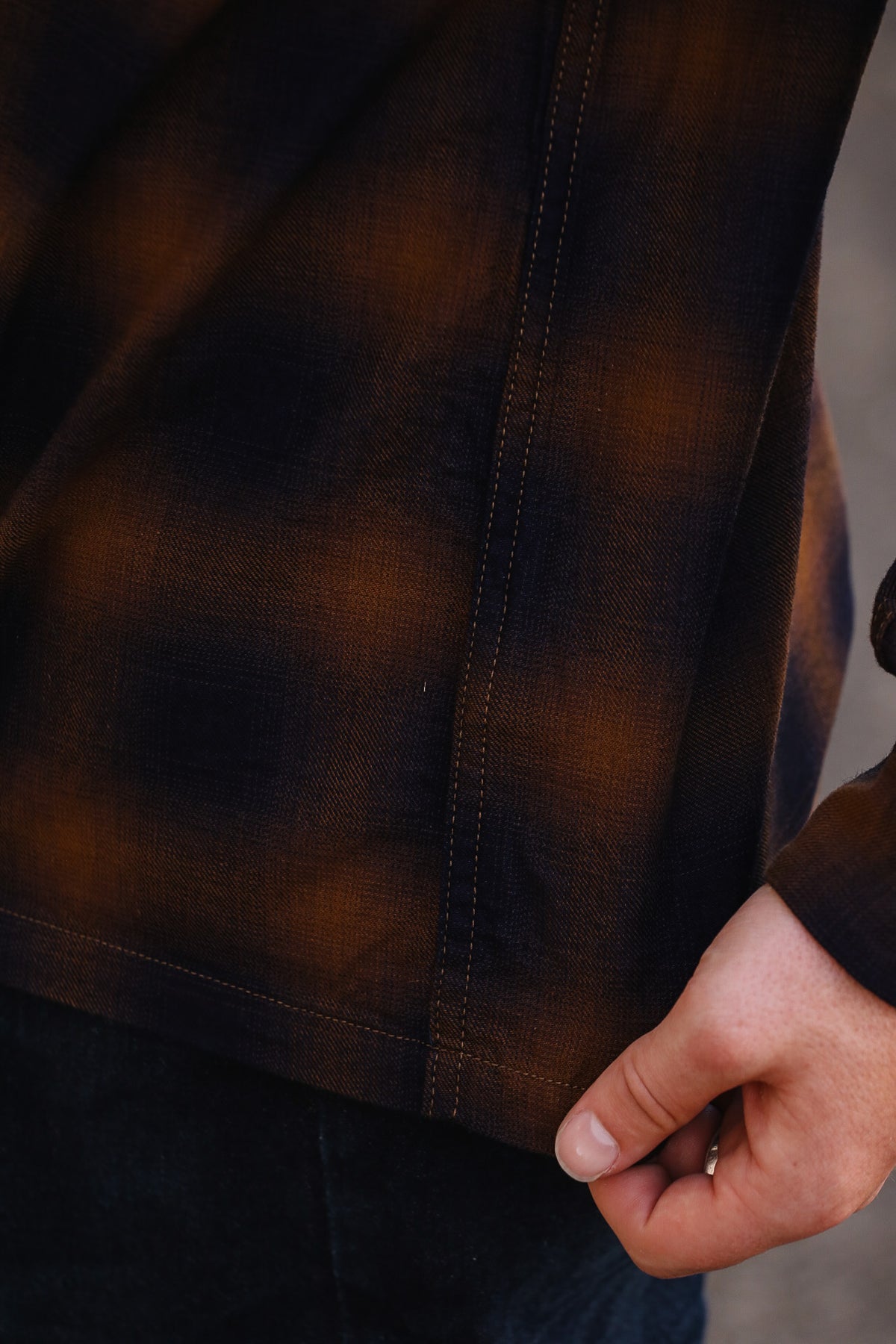 Close-up of a person wearing a dark brown and black plaid flannel shirt, with a hand pulling the hem slightly to show the fabric's soft texture and drape.