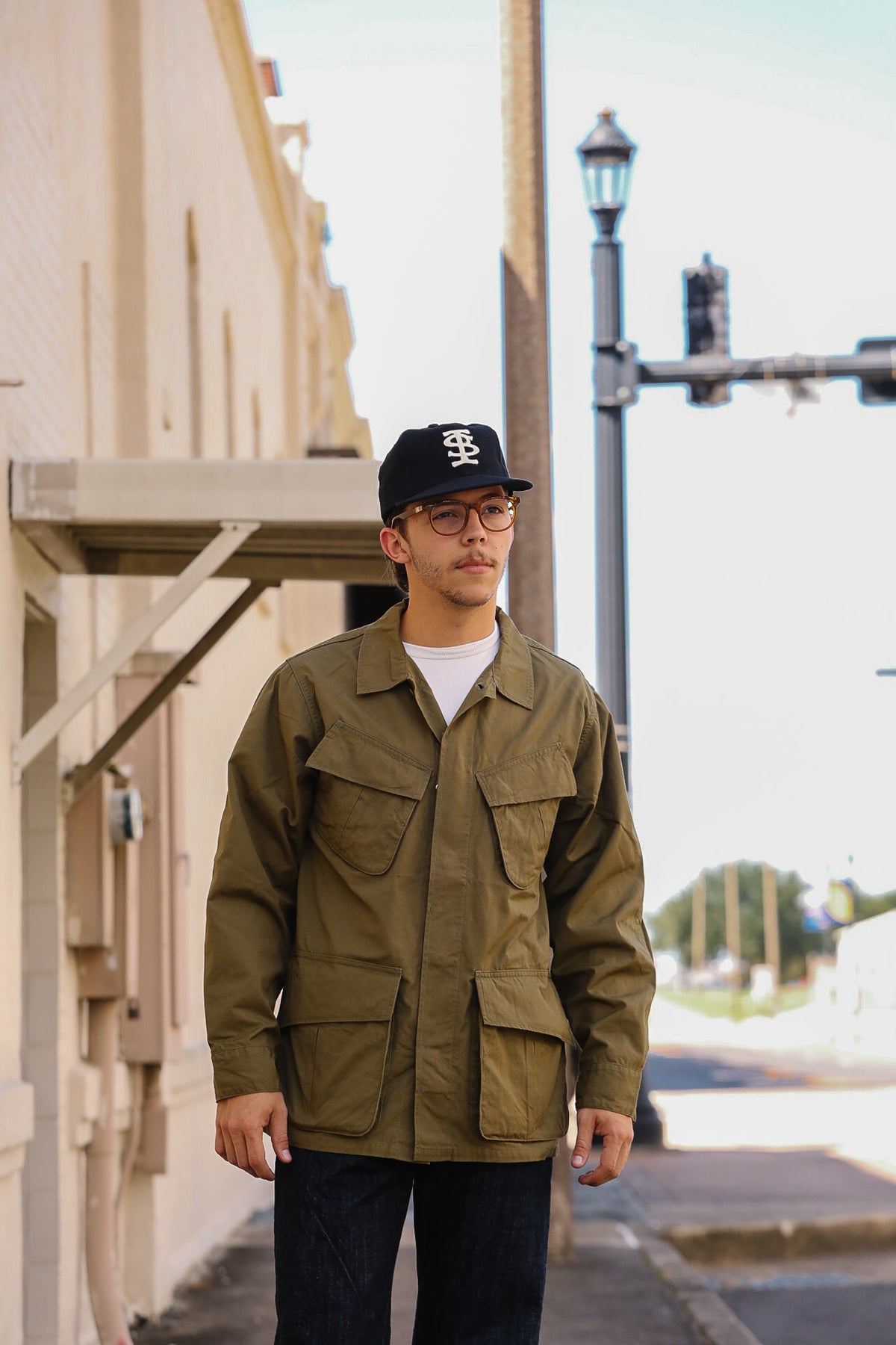 A man wearing an olive green utility jacket, black baseball cap, and glasses standing on a city sidewalk.