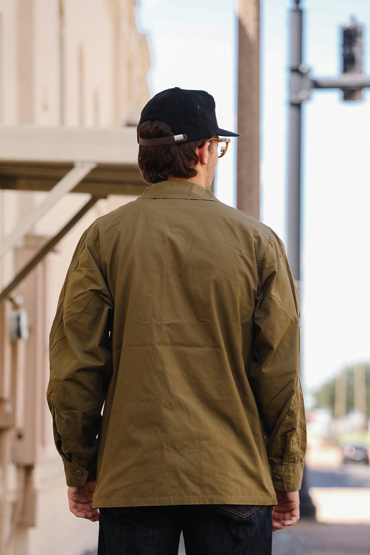 A man viewed from behind wearing an olive green utility jacket, a black baseball cap, and glasses while standing outdoors on a city street.
