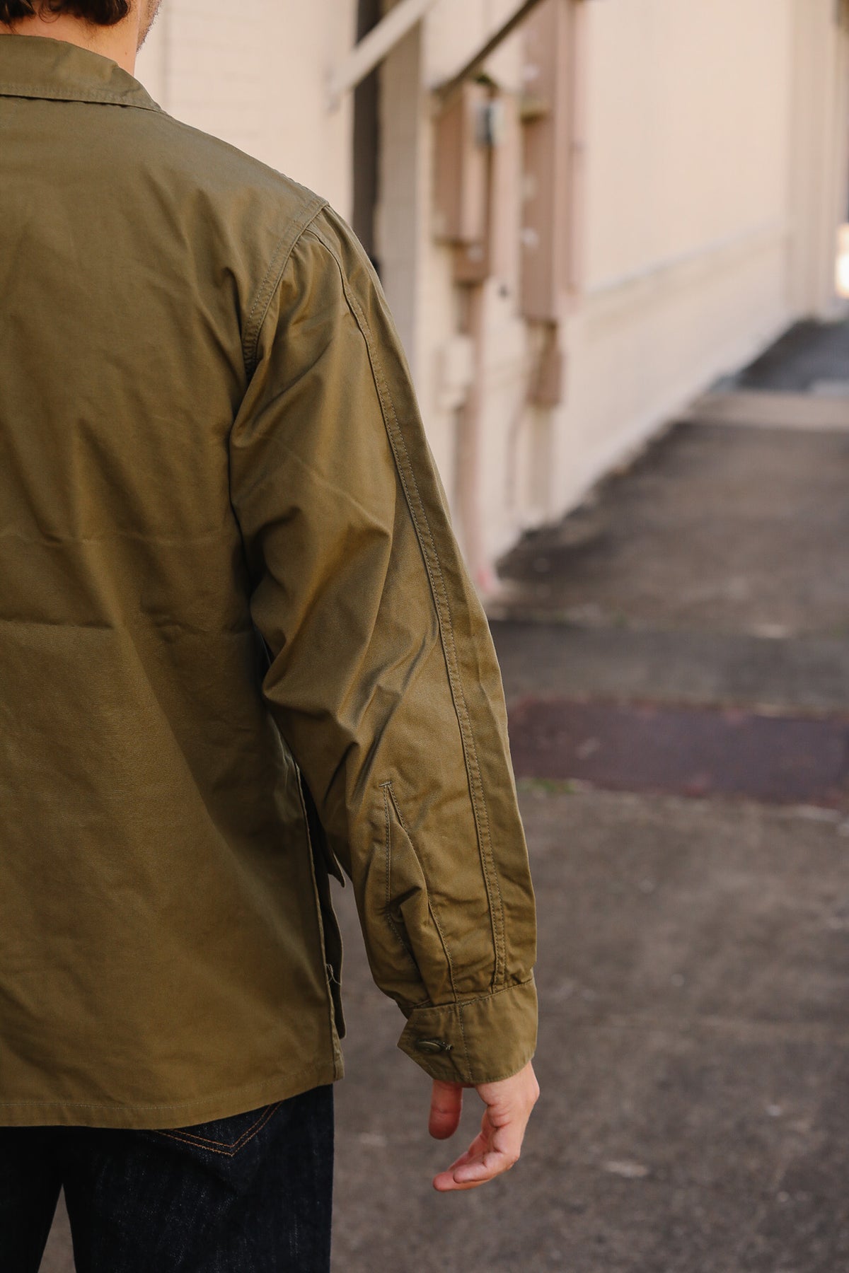 A person viewed from behind wearing an olive green utility jacket and dark jeans, standing on a concrete walkway next to a beige building.