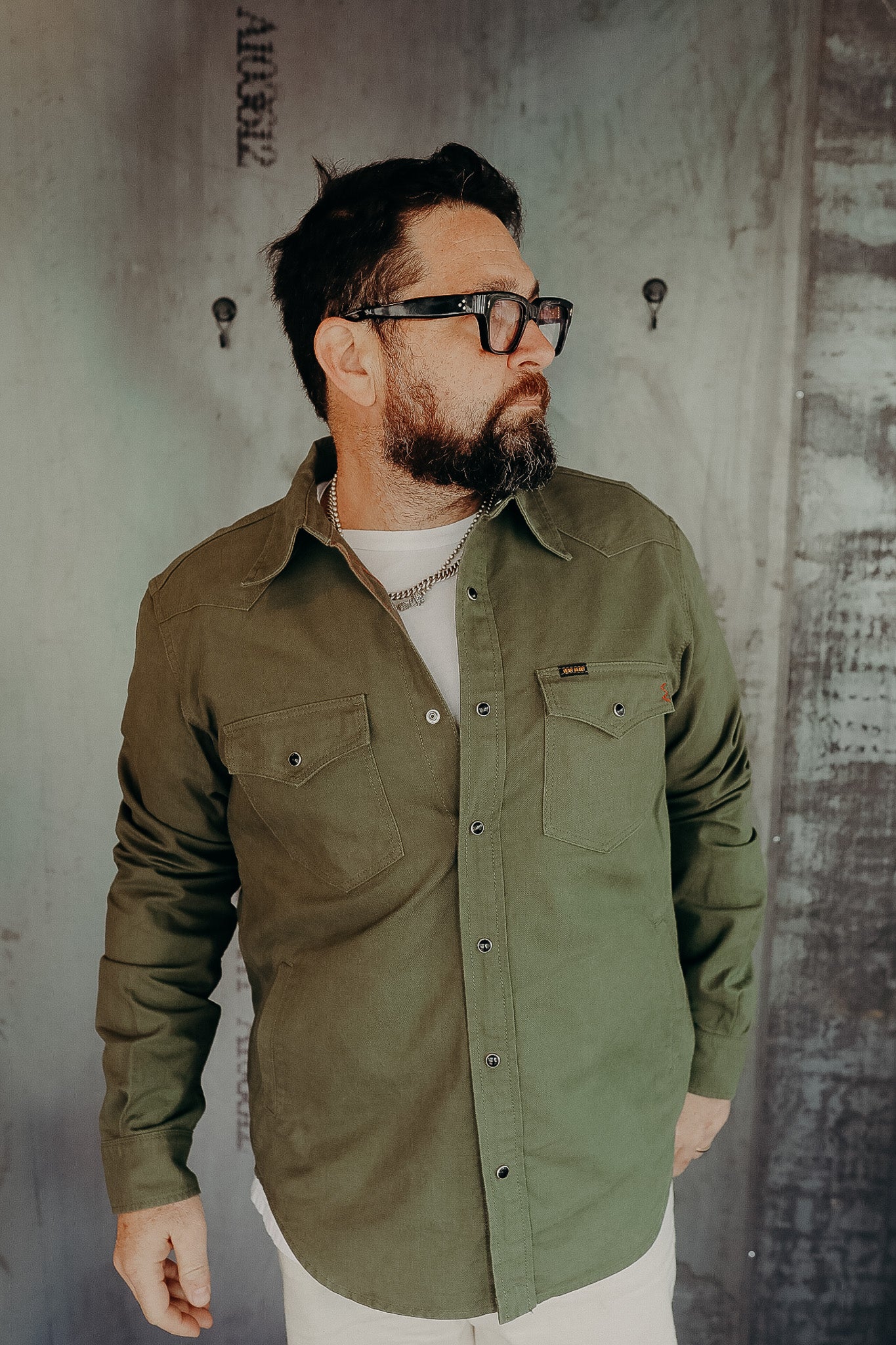 Man wearing olive green button-up work shirt with chest pockets and glasses indoors