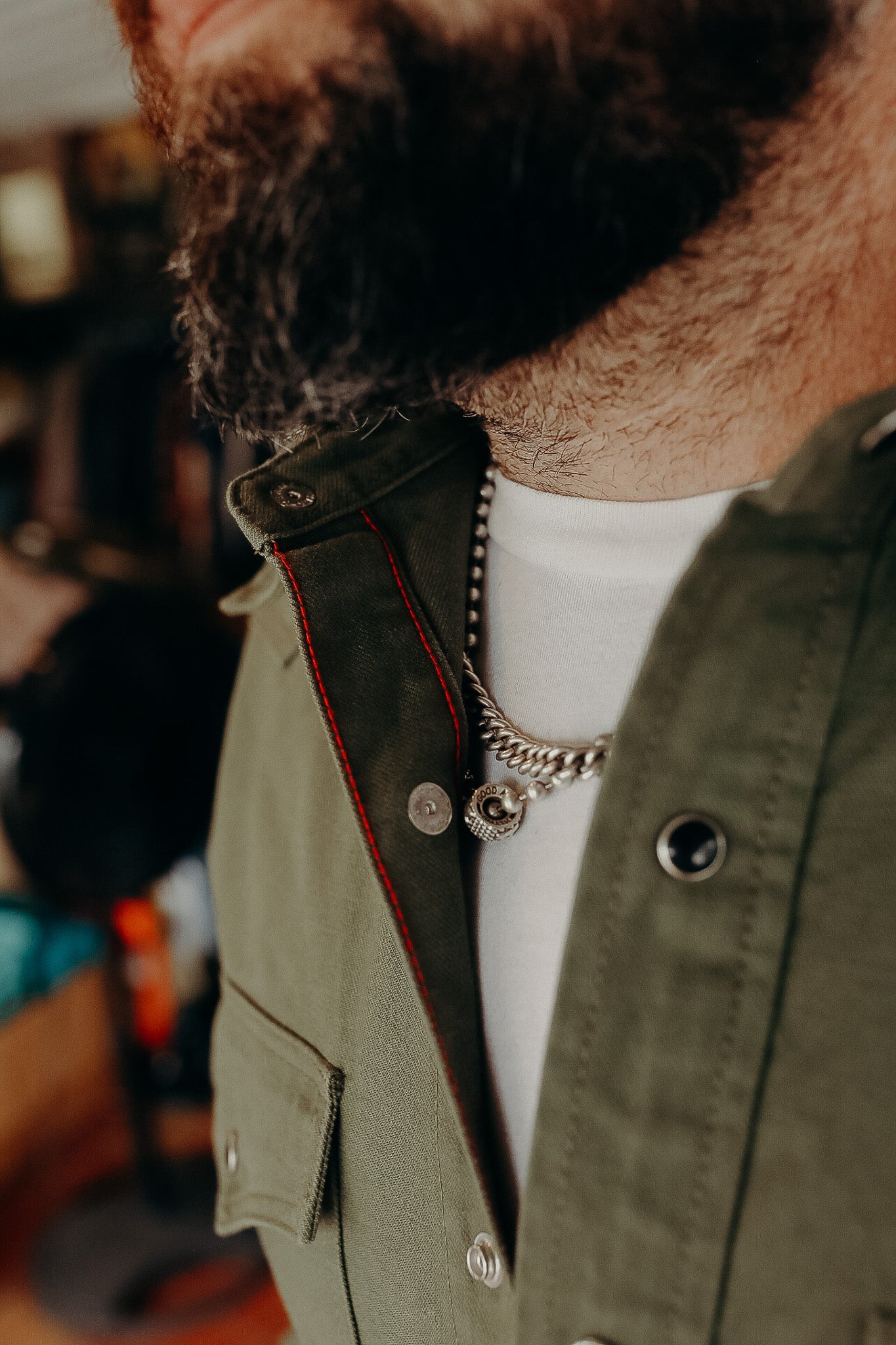 Close-up of a bearded man wearing an olive green jacket with red stitching and a silver chain necklace over a white shirt