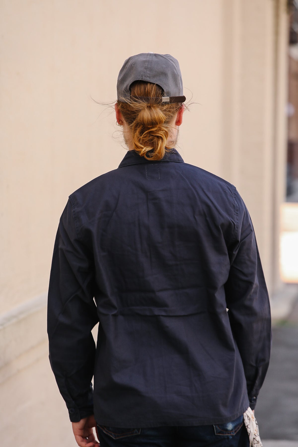 A person viewed from behind wearing a dark navy blue long-sleeve utility-style jacket and a gray baseball cap, standing outdoors against a light beige wall.