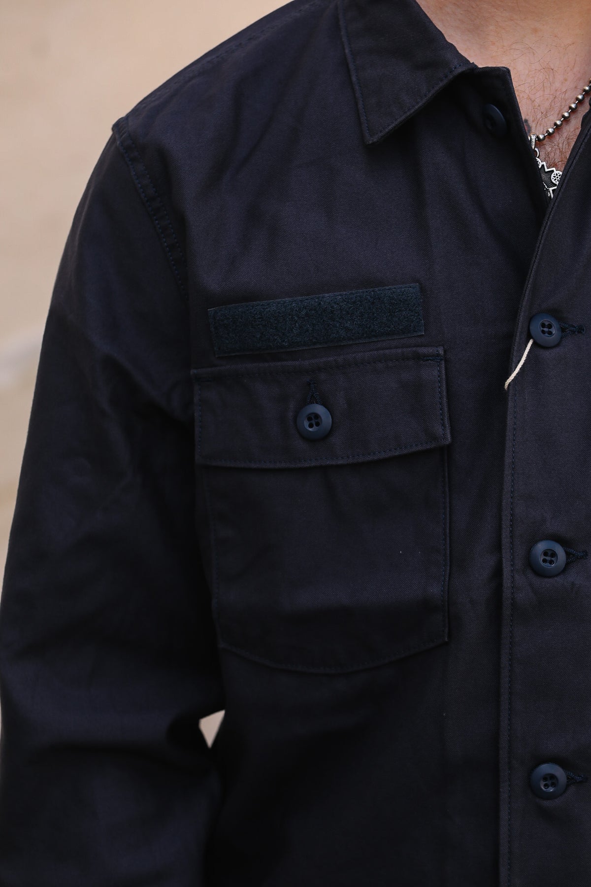 Close-up of a person wearing a dark navy or near-black button-front overshirt with a chest pocket and a dark rectangular patch on the pocket flap.
