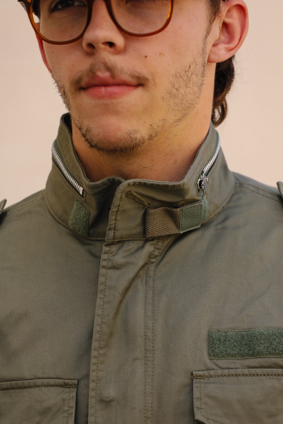 Close-up of a person wearing an olive green military-style jacket with a high collar, zipper details, and a patch pocket, paired with round tortoiseshell glasses against a plain beige background.