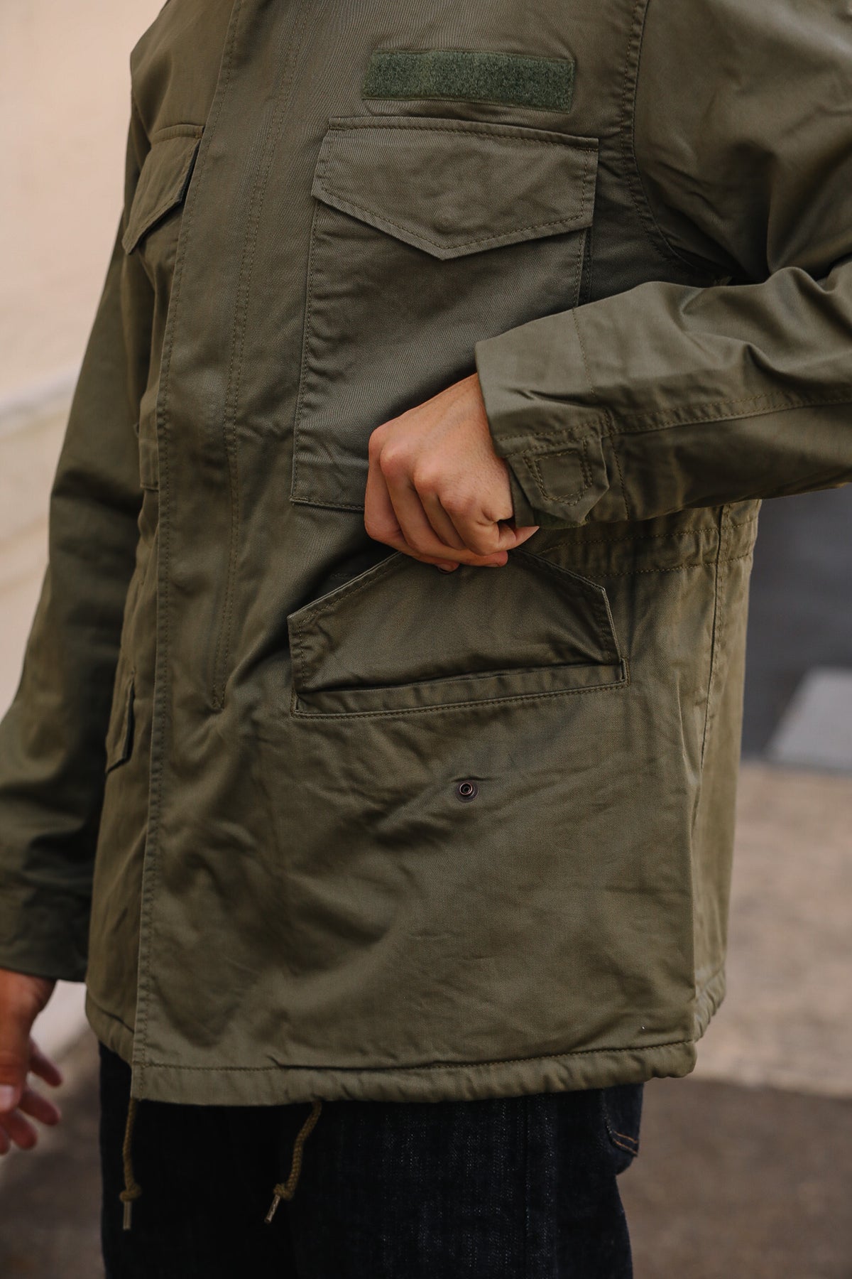 Close-up of a person wearing an olive green military-style jacket with multiple flap pockets and a hand tucked into a front pocket.