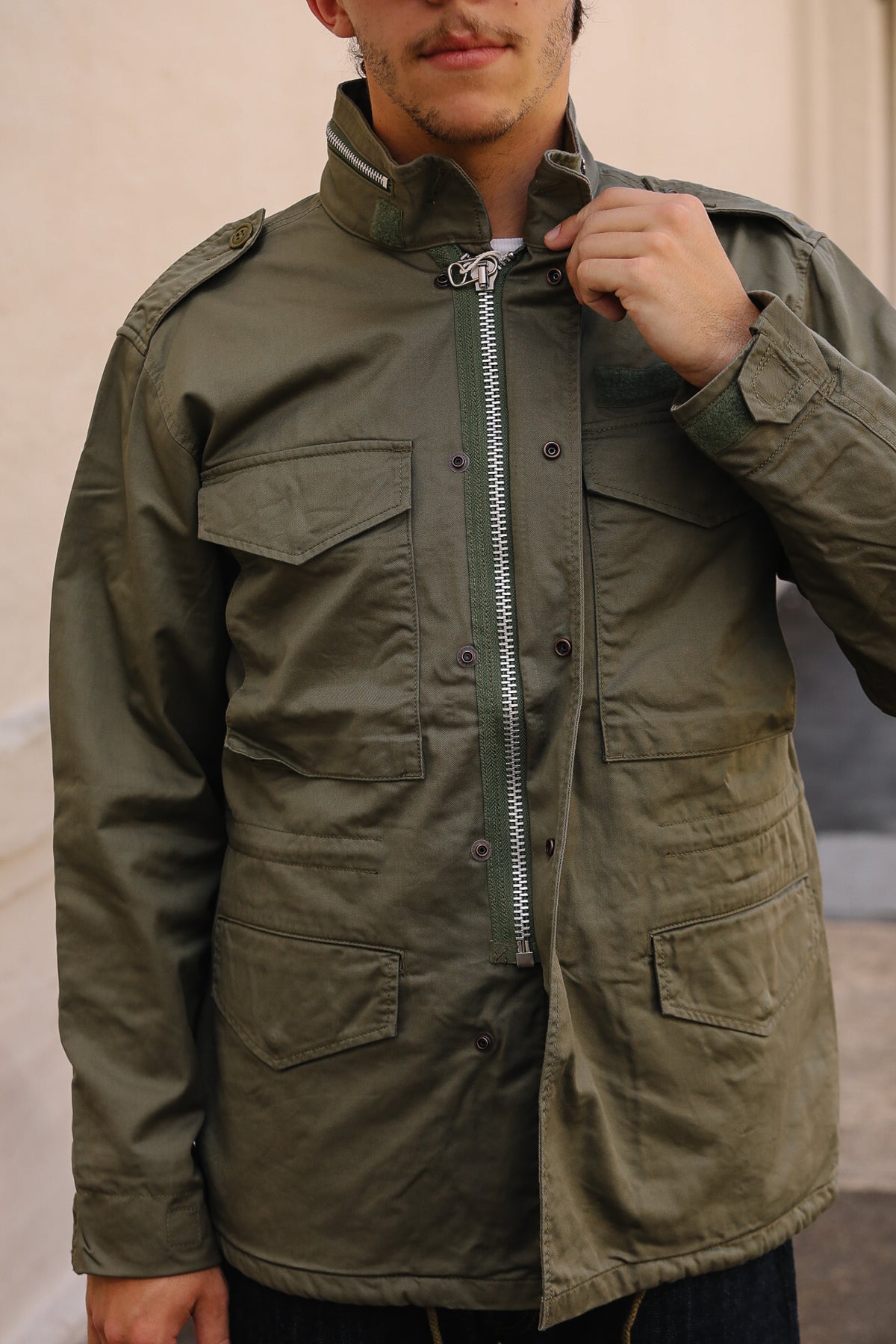 A man wearing an olive green military-style field jacket with a high collar, front zipper, and multiple flap pockets, shown from the chin down against a neutral outdoor background.