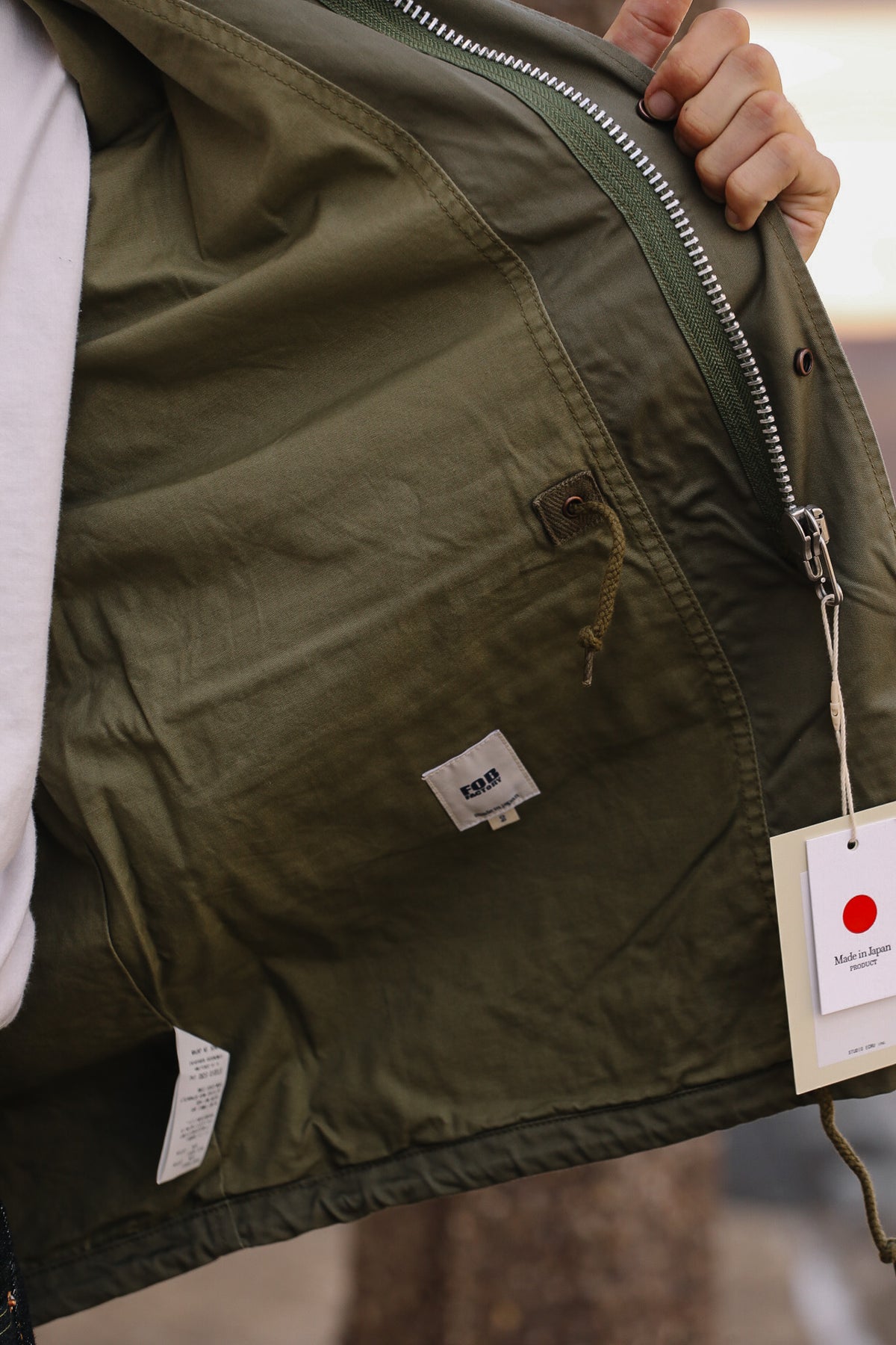 Close-up of a person holding open an olive green jacket, showing the interior lining, zipper, pocket detail, and hanging product tags.