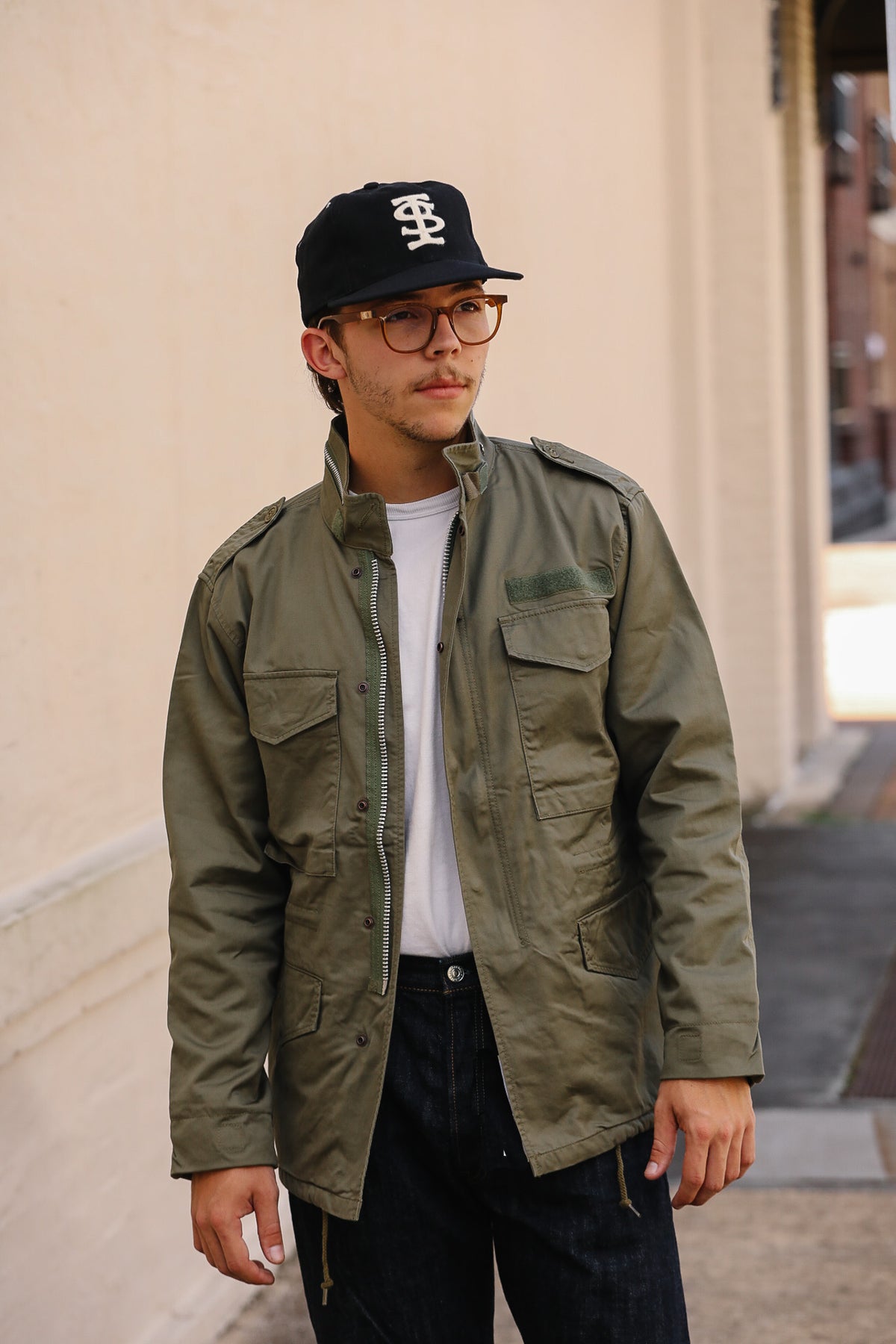 A man wearing an olive green military-style field jacket over a white t-shirt, dark jeans, and a black baseball cap with a white logo, standing outdoors against a beige wall.