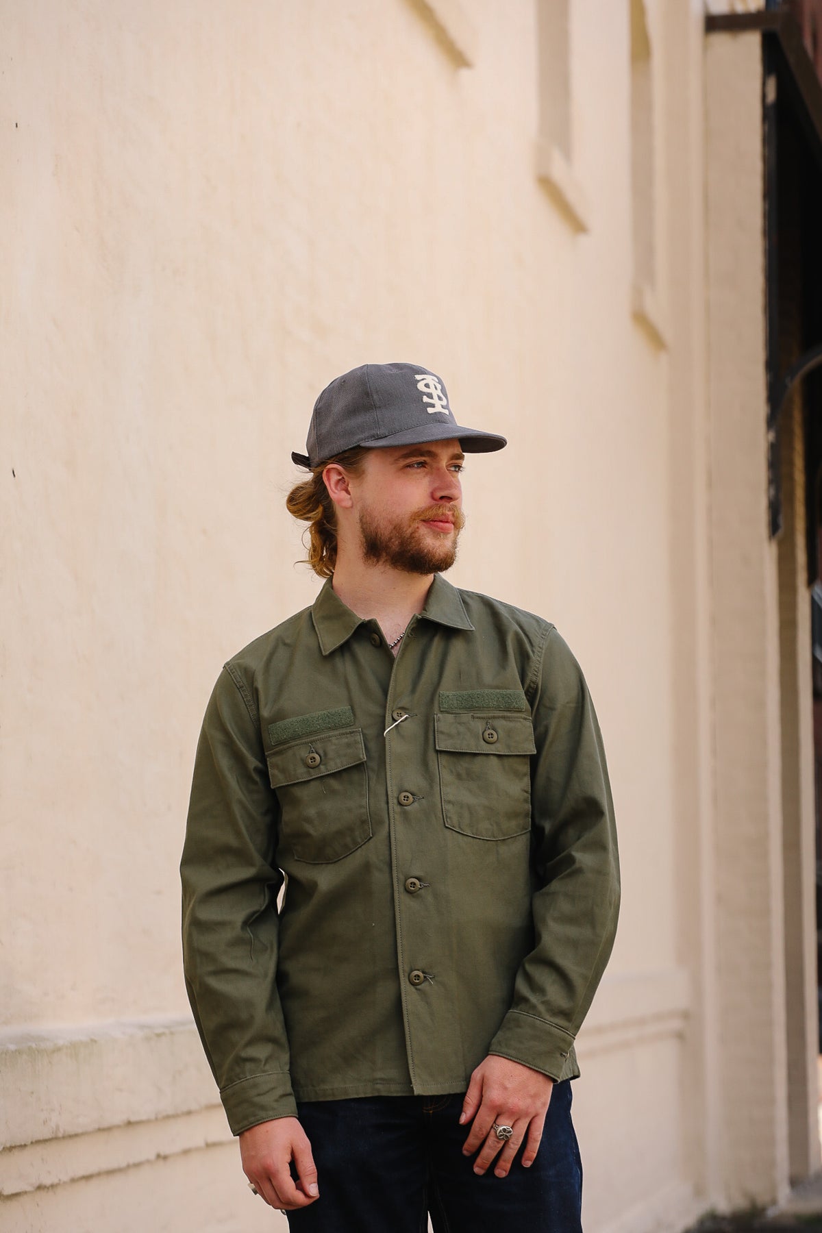 A man wearing an olive green utility shirt jacket and a dark gray baseball cap with a white logo, standing against a beige stucco building wall.