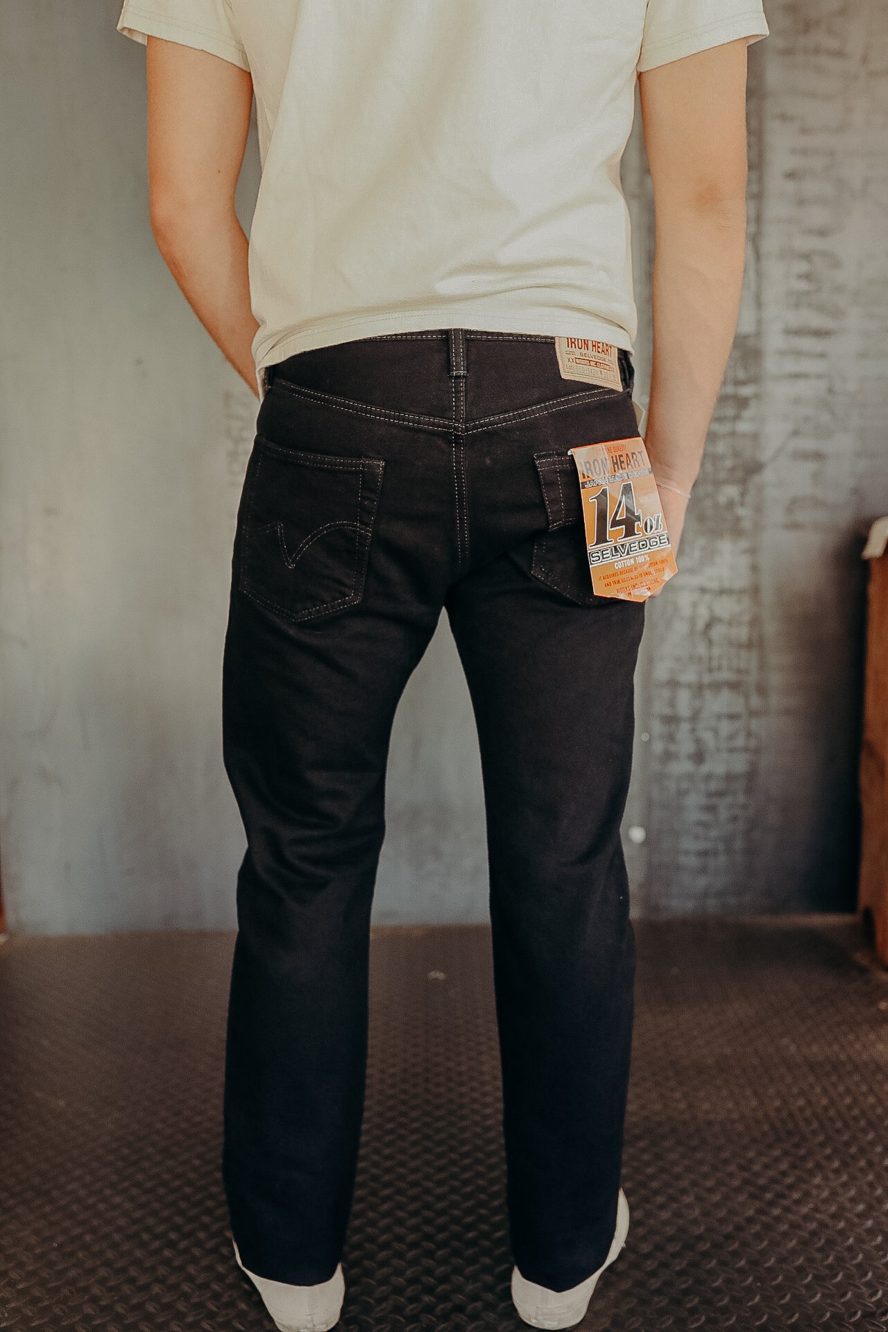 Back view of person wearing black Iron Heart 14oz selvedge denim jeans with label tag, casual beige t-shirt