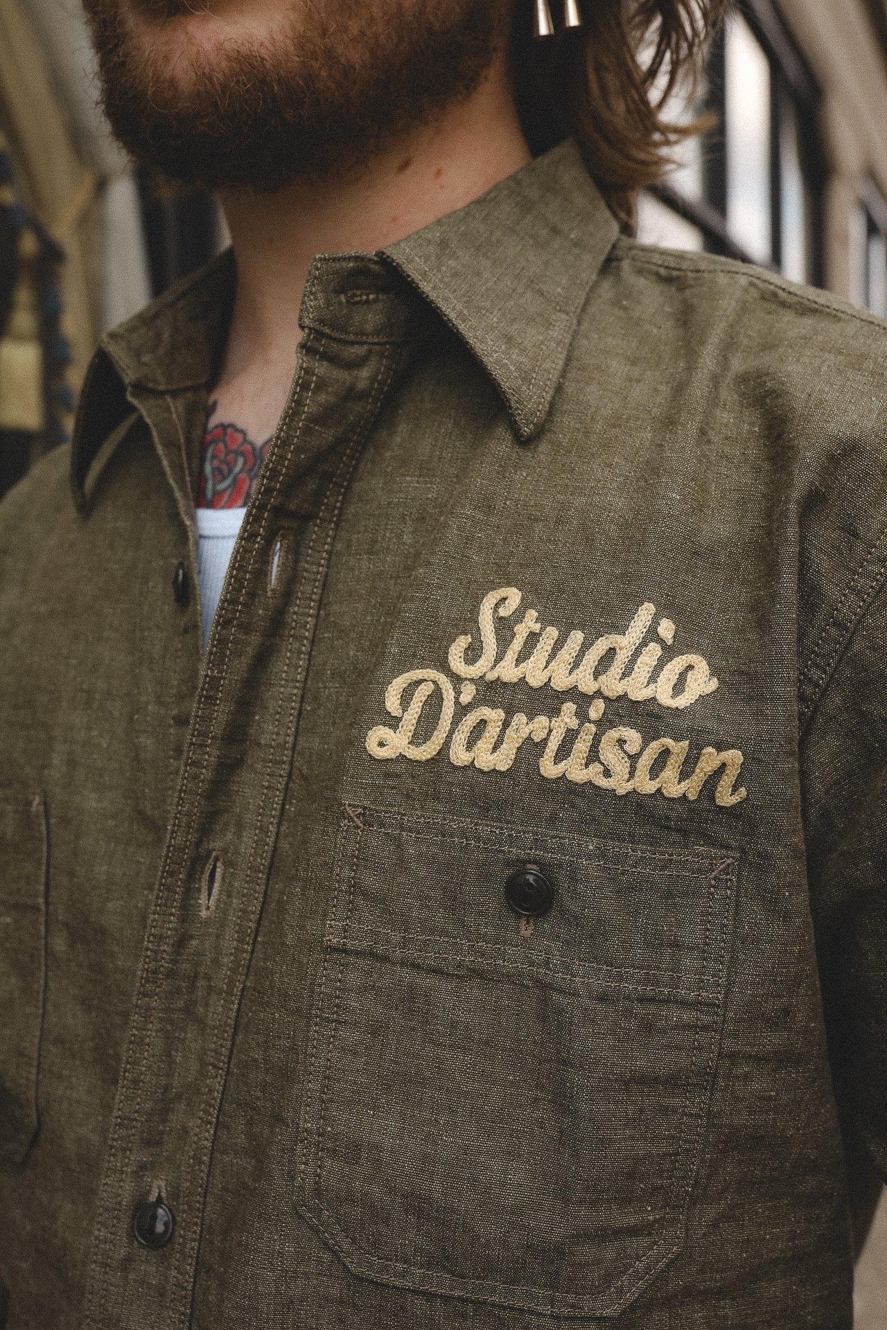 Close-up of man wearing olive green shirt with embroidered 'Studio D'artisan' logo and chest pocket