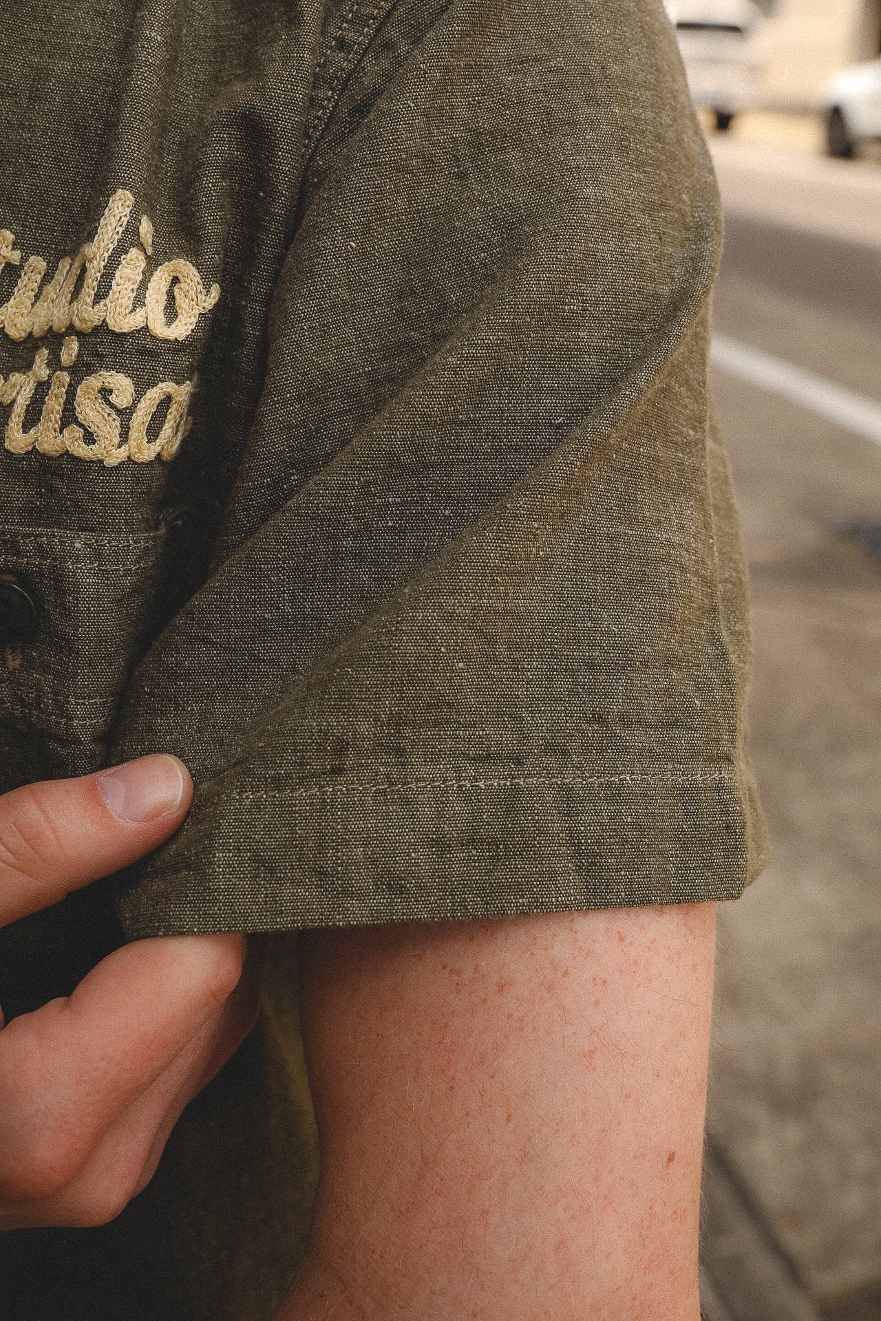 Close-up of a person's hand holding the cuff of an olive green short sleeve work shirt embroidered with 'Studio Artisans'