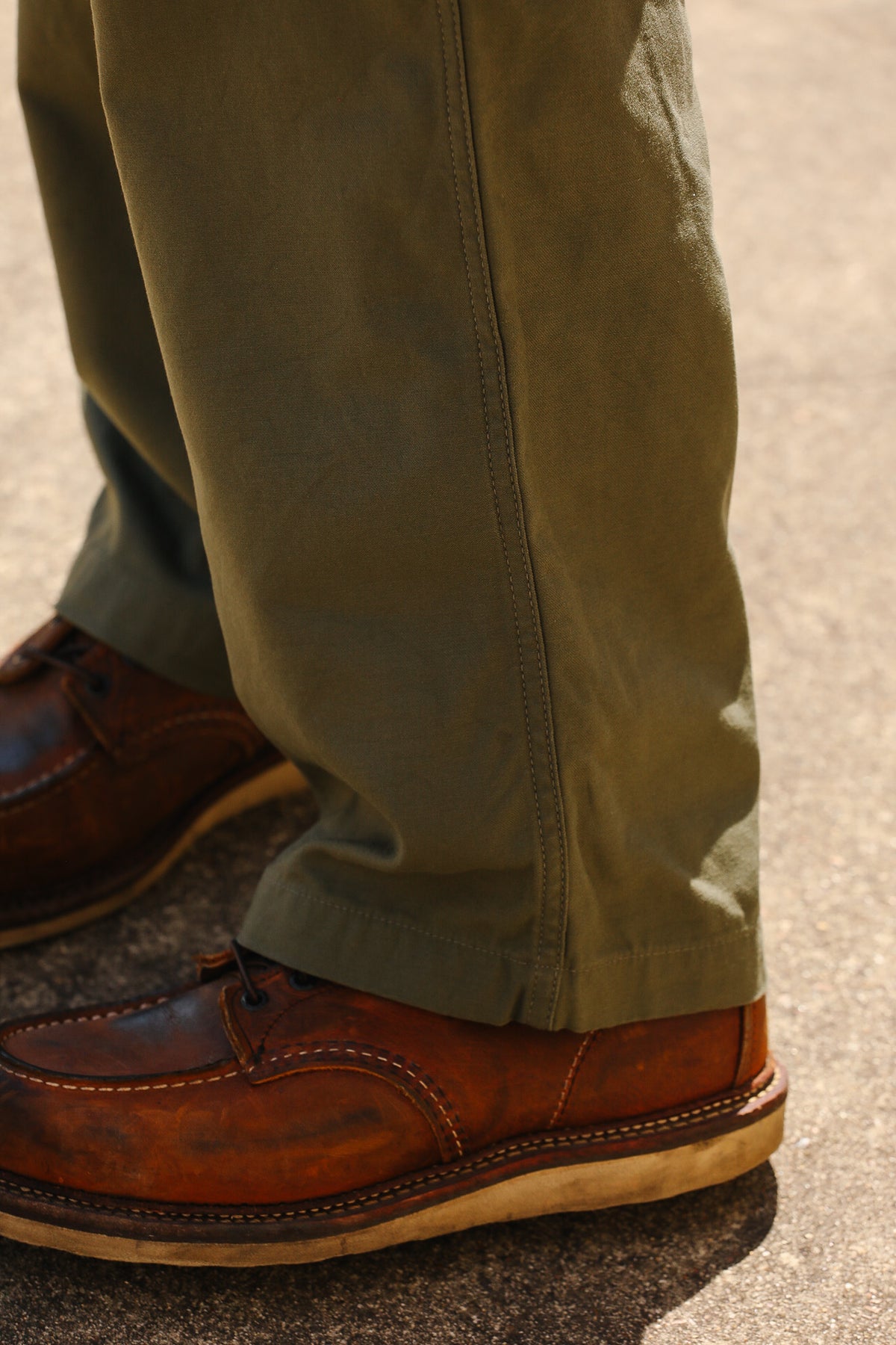 Close-up of a person's legs wearing olive green pants and brown leather boat shoes on a gray asphalt surface.