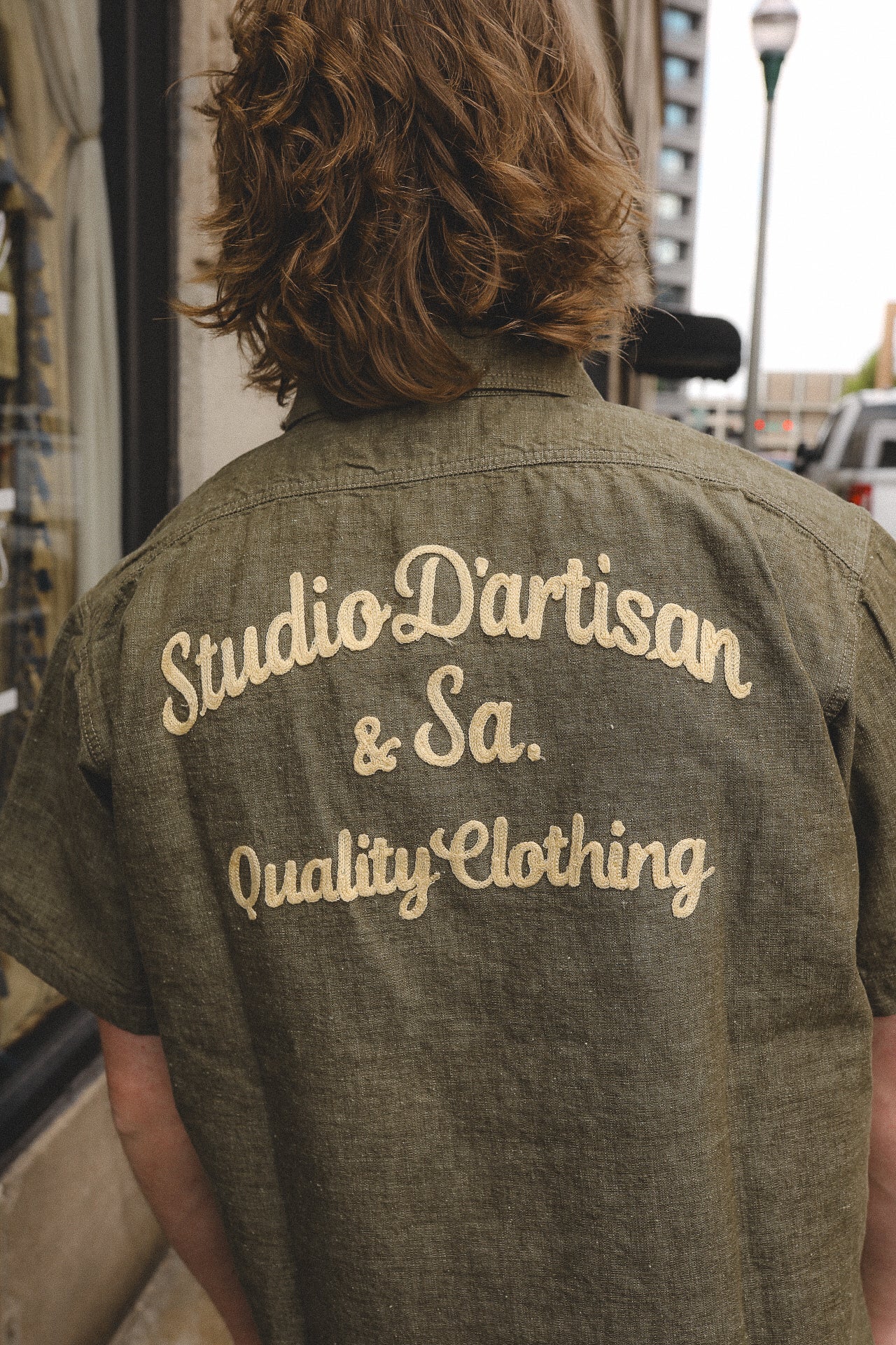 Back view of a person wearing an olive green shirt embroidered with 'Studio D'artisan &amp; Sa. Quality Clothing' outdoors in a city street