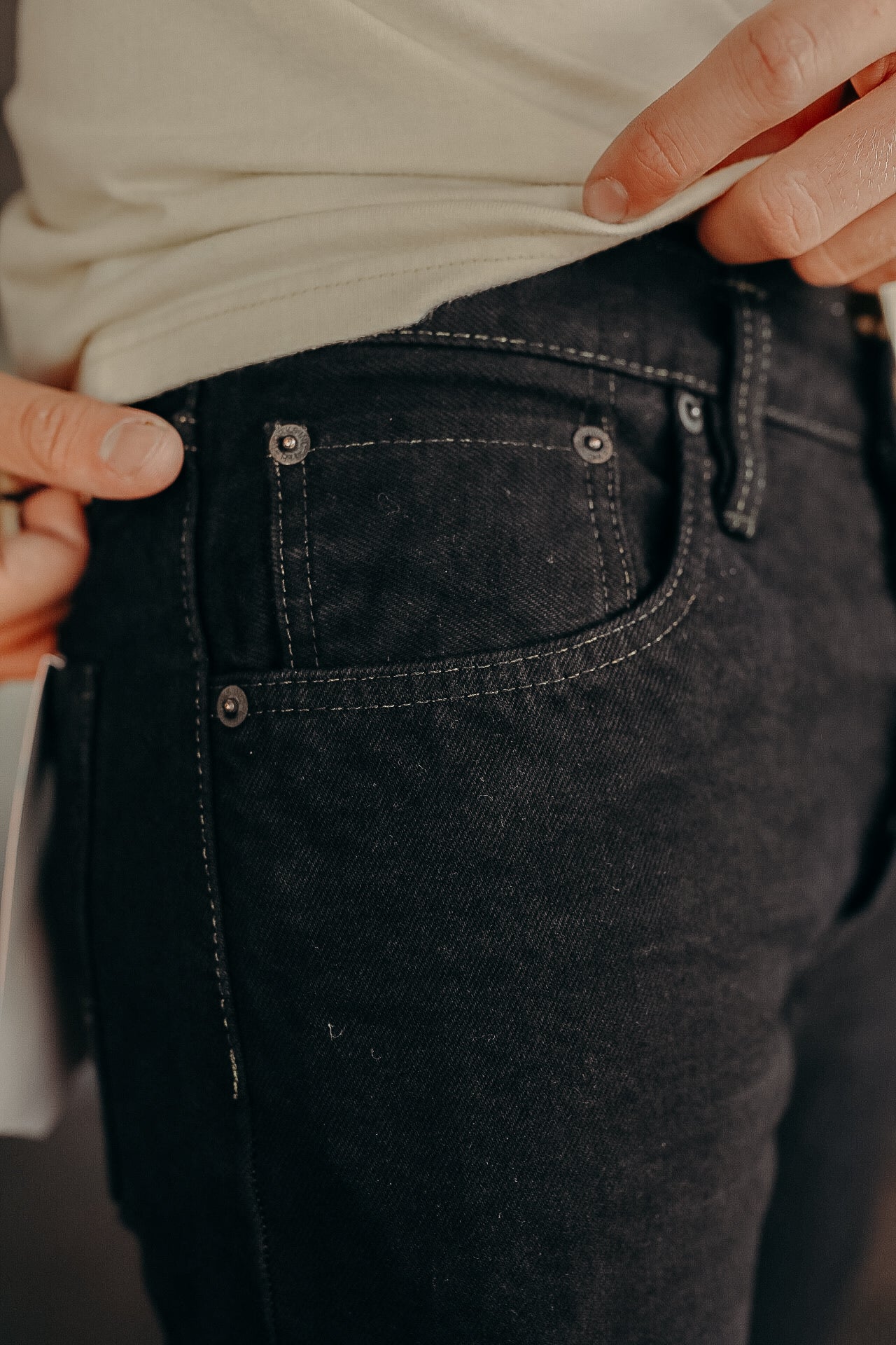 Close-up of a person wearing black denim jeans with detailed stitching and silver rivets, lifting beige shirt